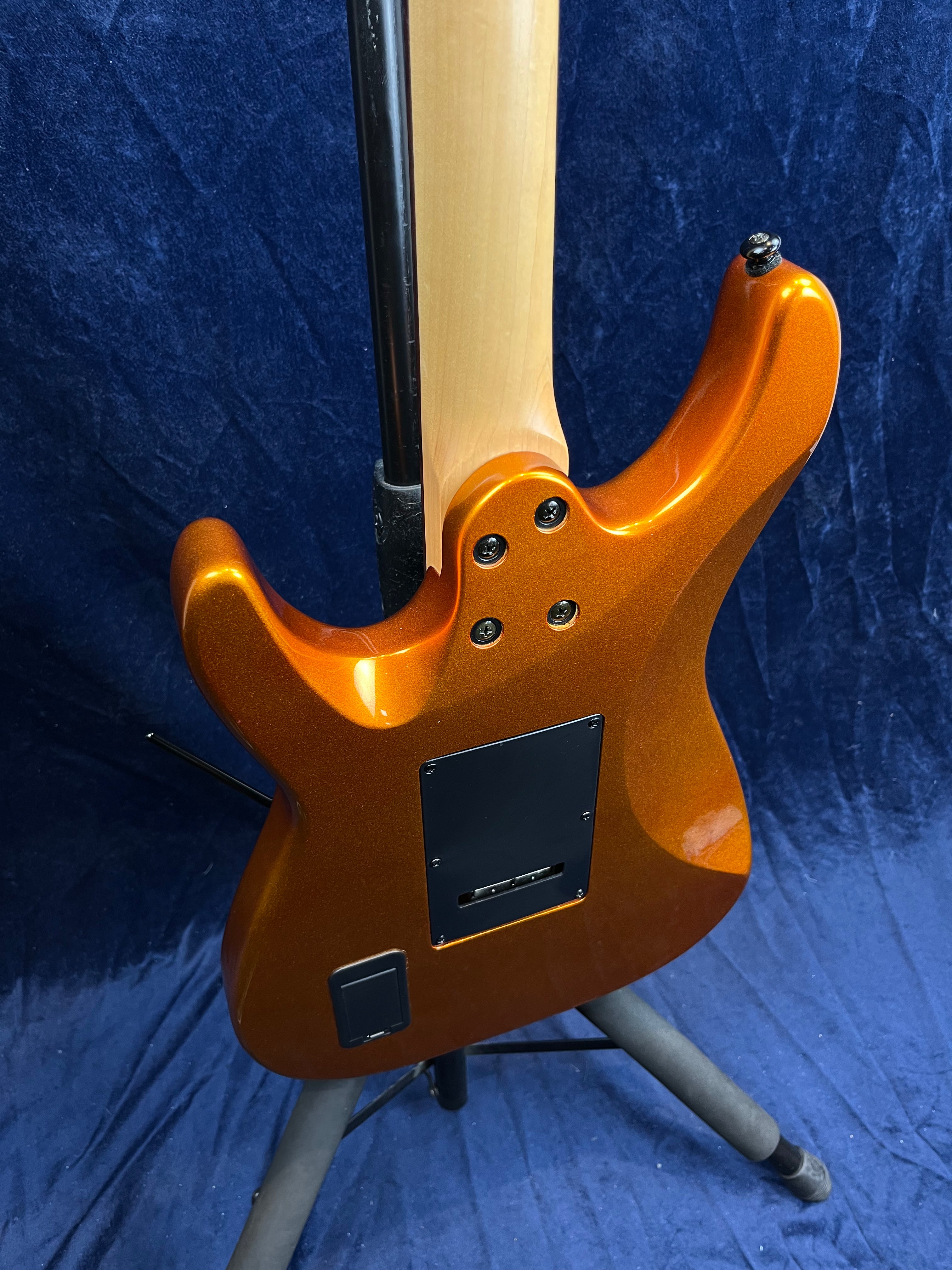 Electric guitar in metallic orange, shown from the back with a natural wood neck, two black back plates, and mounted on a stand against a blue background.