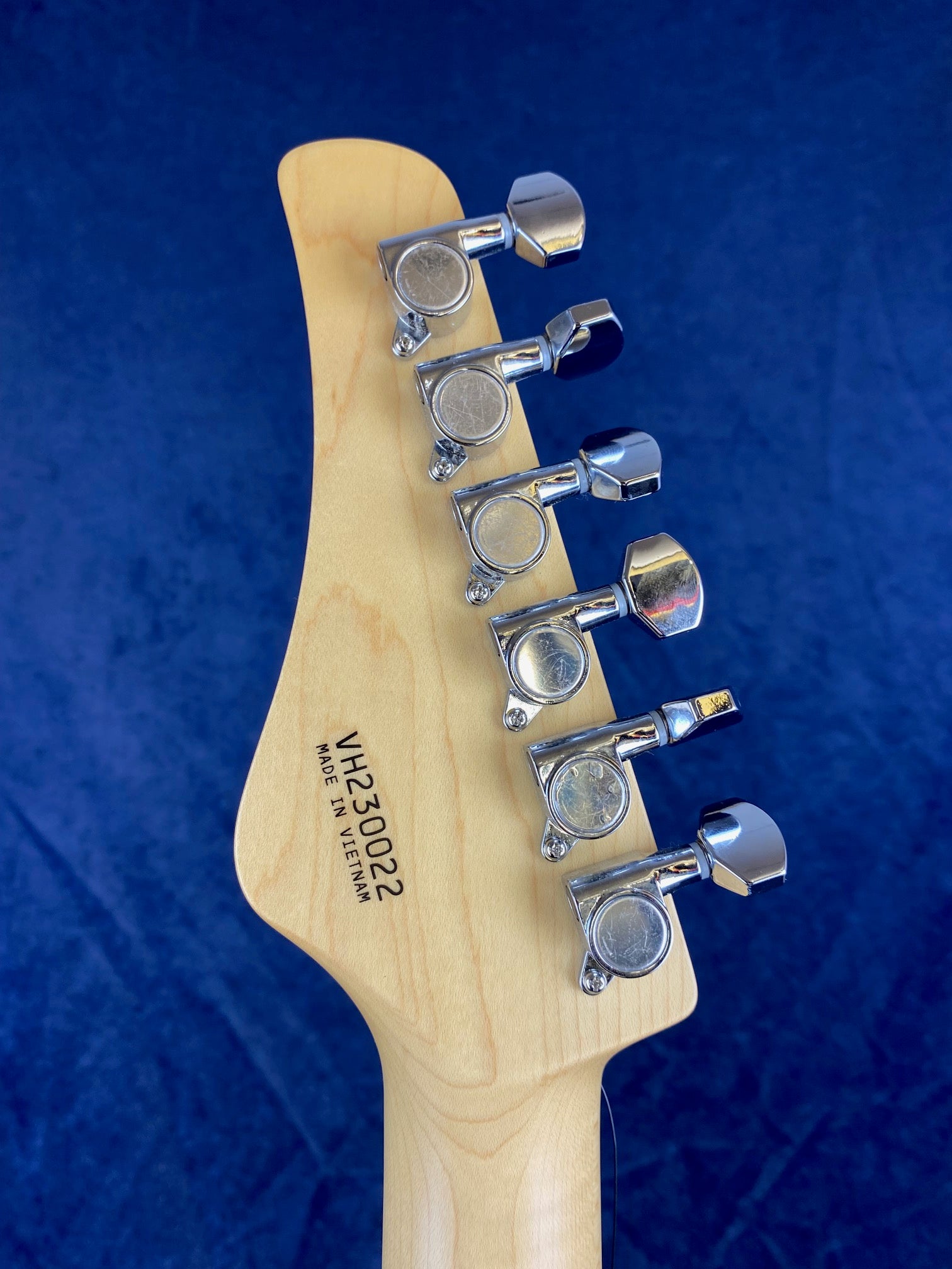 Six chrome tuning pegs on a light wood electric guitar headstock, serial number VH230022, made in Vietnam, against a blue background.