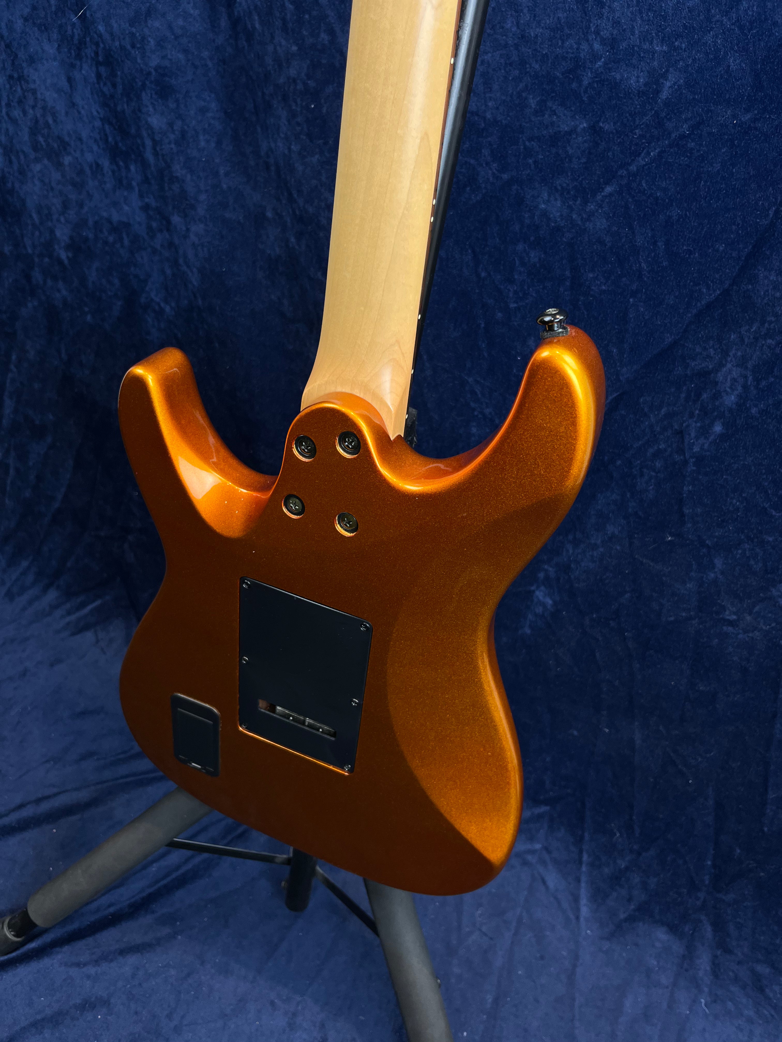 Metallic orange electric guitar shown from the back, with a natural wood neck and black plastic covers on the body, standing on a guitar stand.