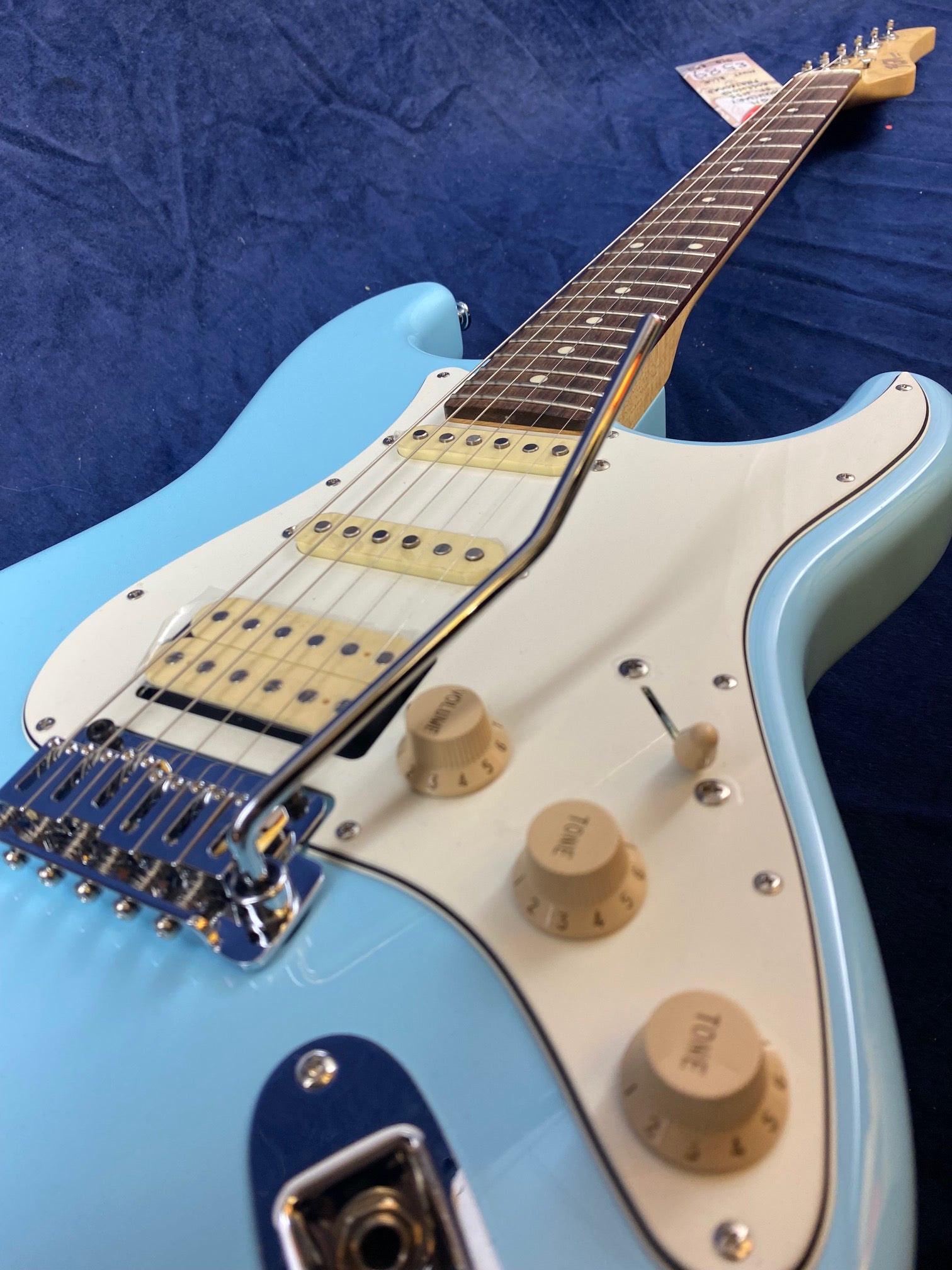 Light blue electric guitar with a white pickguard, three single-coil pickups, three control knobs, and a tremolo arm, lying on a dark blue surface.