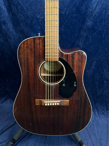 Dark wood acoustic guitar with a cutaway design, black pickguard, and six strings, standing upright against a dark blue background.