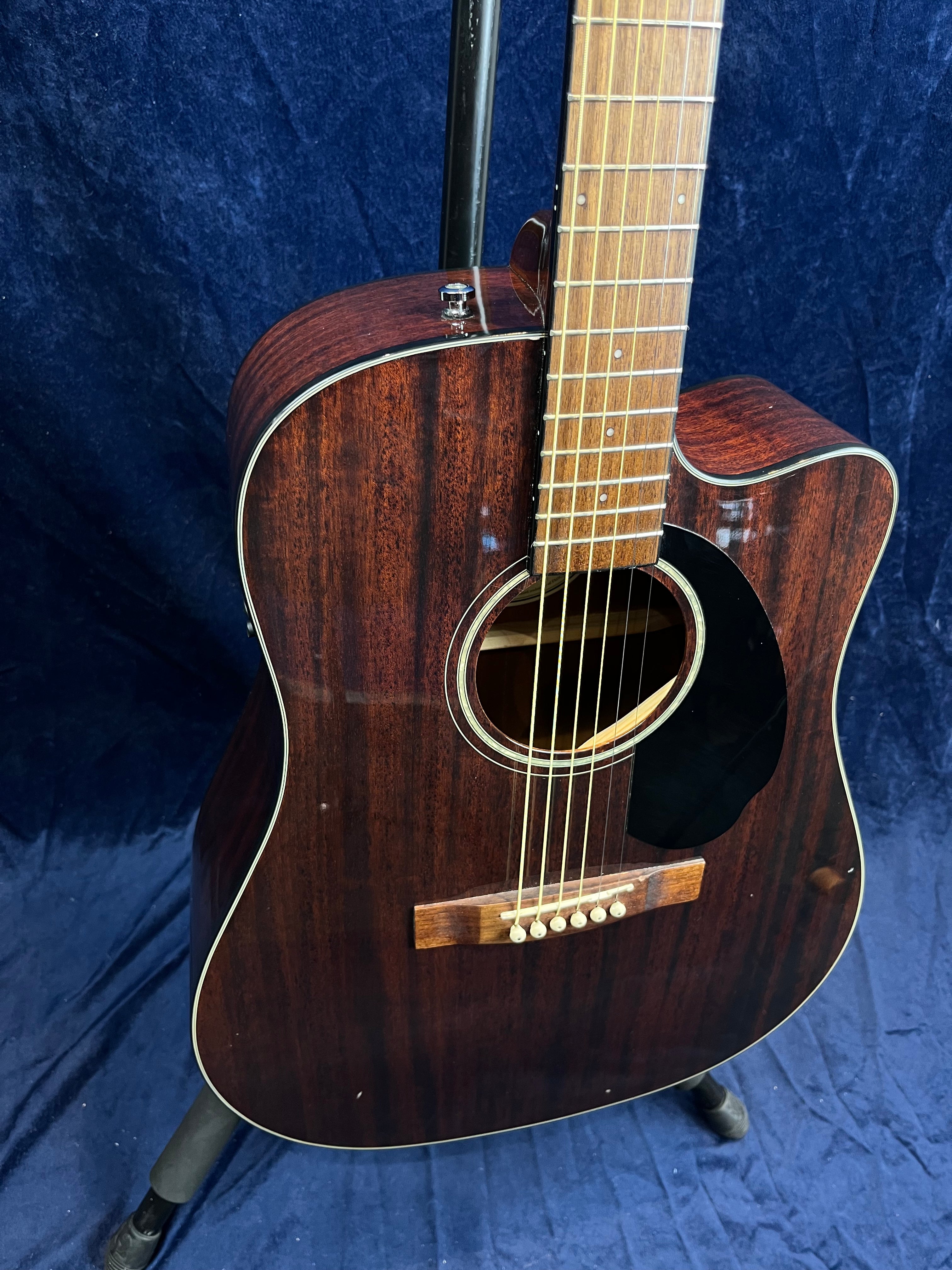 Dark wood acoustic guitar with a cutaway body, six strings, white dot fret markers, and a black pickguard, leaning on a stand against a dark background.