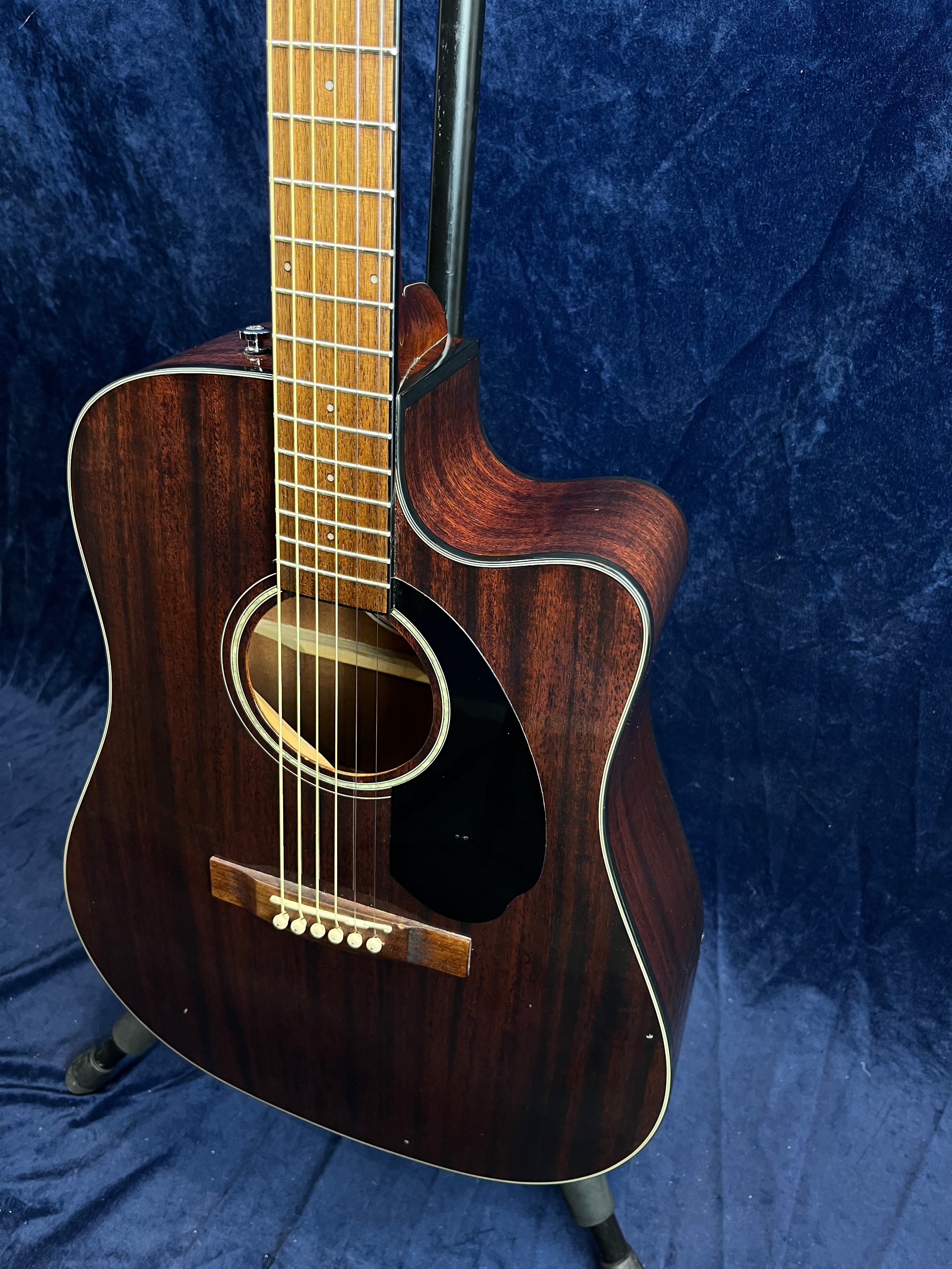 Dark wood acoustic guitar with a cutaway body, light wood fretboard, and six strings, displayed upright against a dark blue textured background.