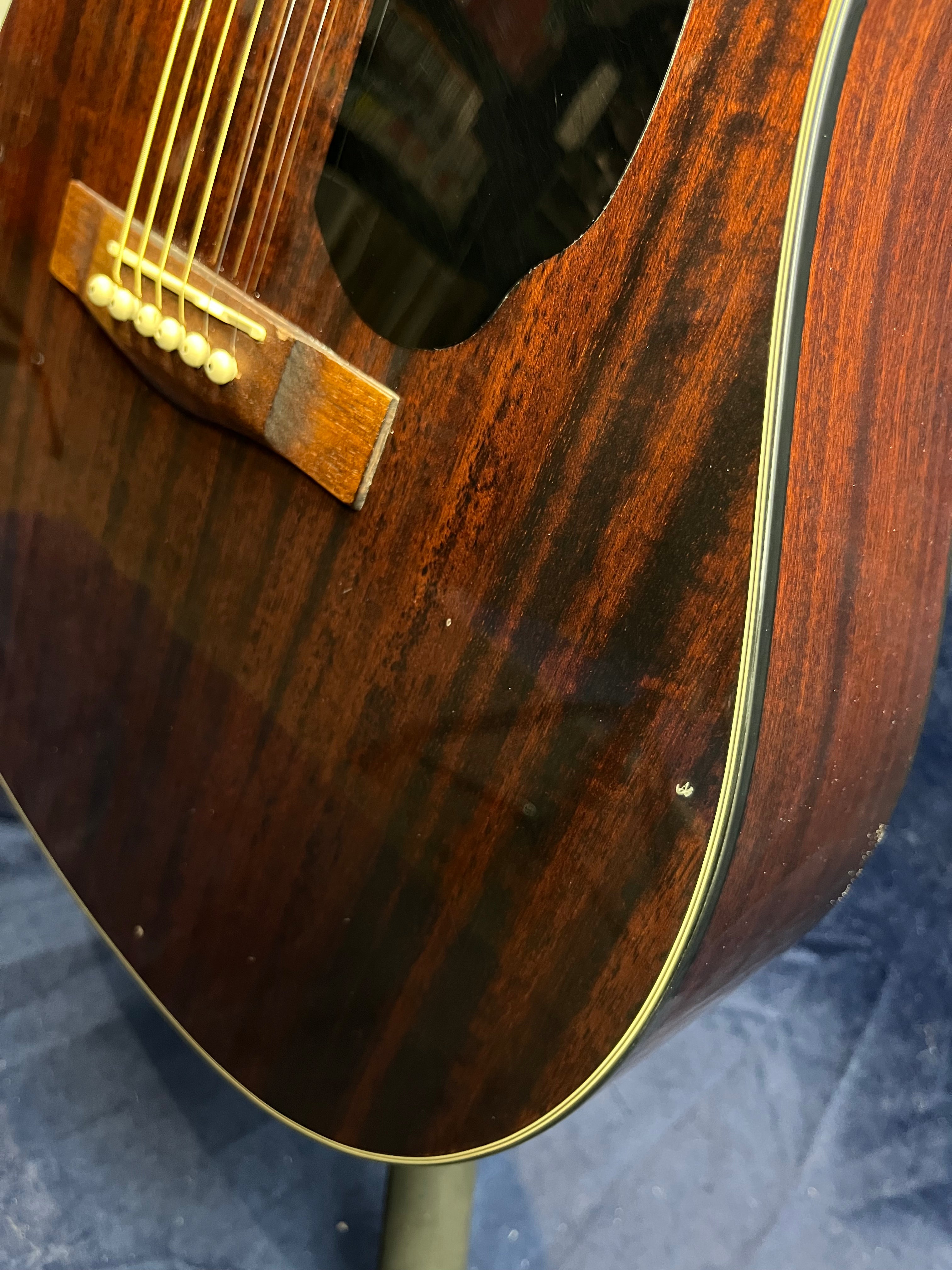 Close-up of an acoustic guitar’s body showing dark wood grain, cream binding, bridge, six strings, and minor surface dings.