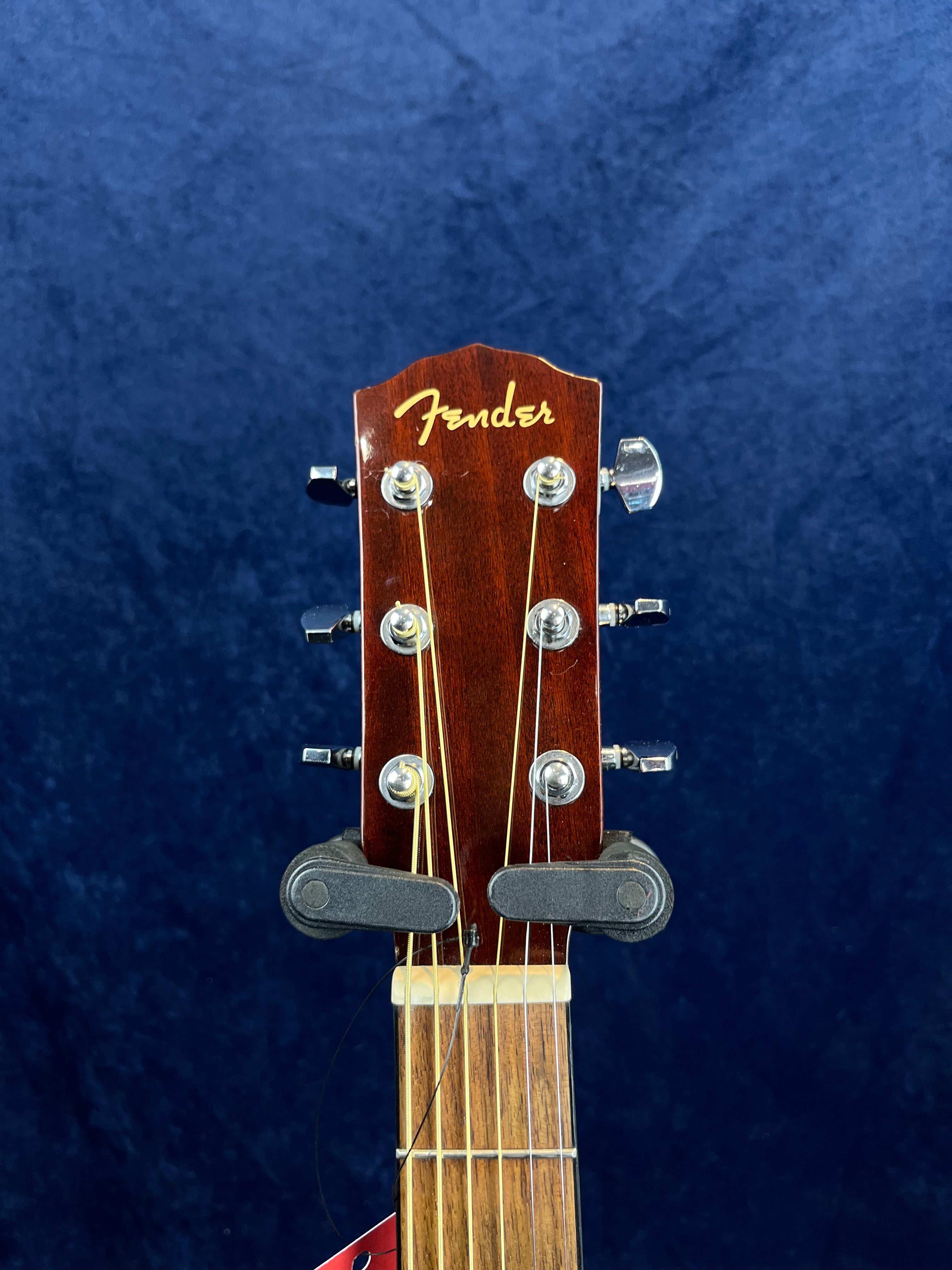 Wooden Fender acoustic guitar headstock with six metal tuning pegs, gold Fender logo, and partial view of frets against a dark blue background.