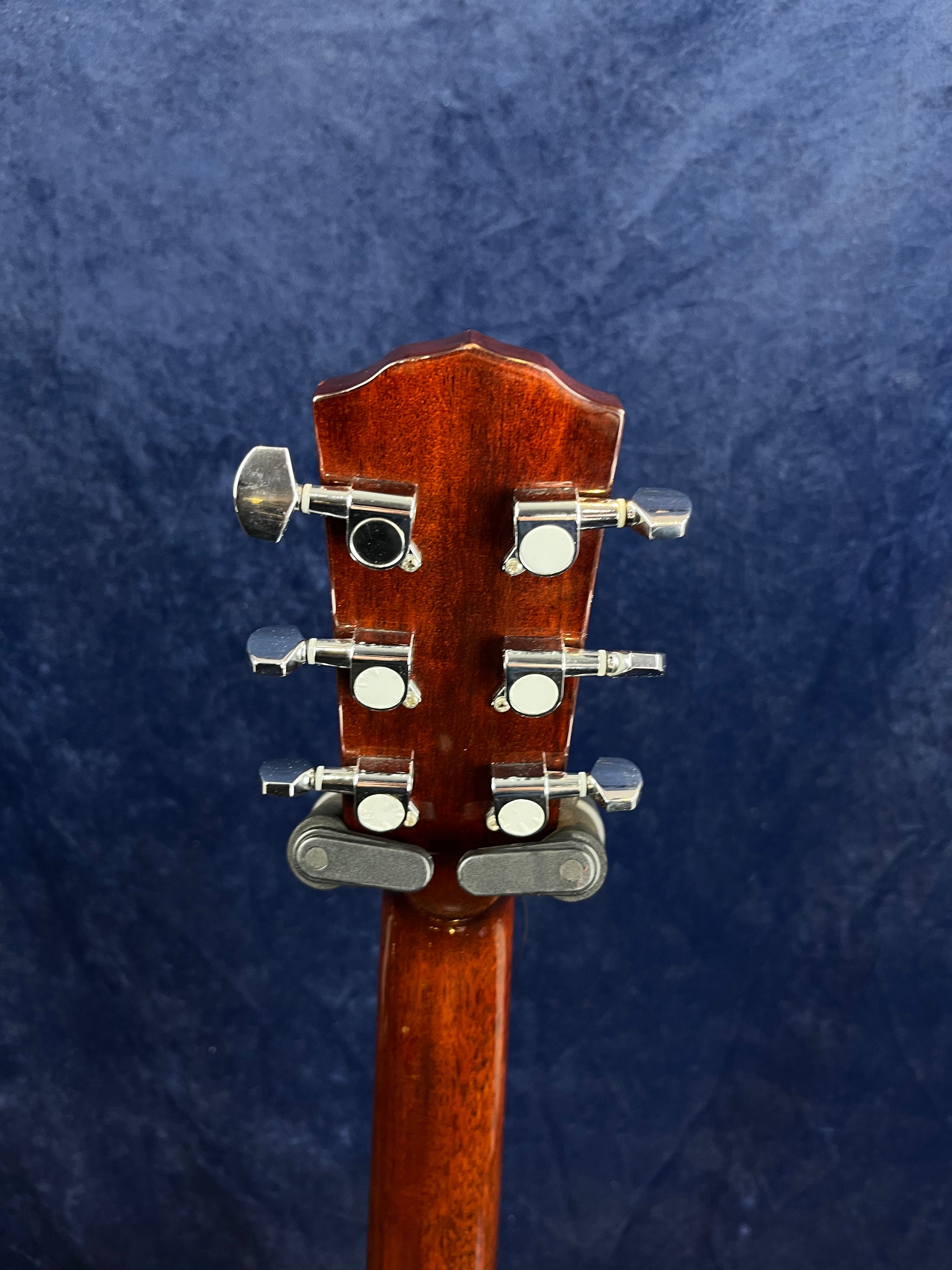 Rear view of an acoustic guitar headstock showing six silver tuning pegs on a polished brown wood neck against a dark blue background.