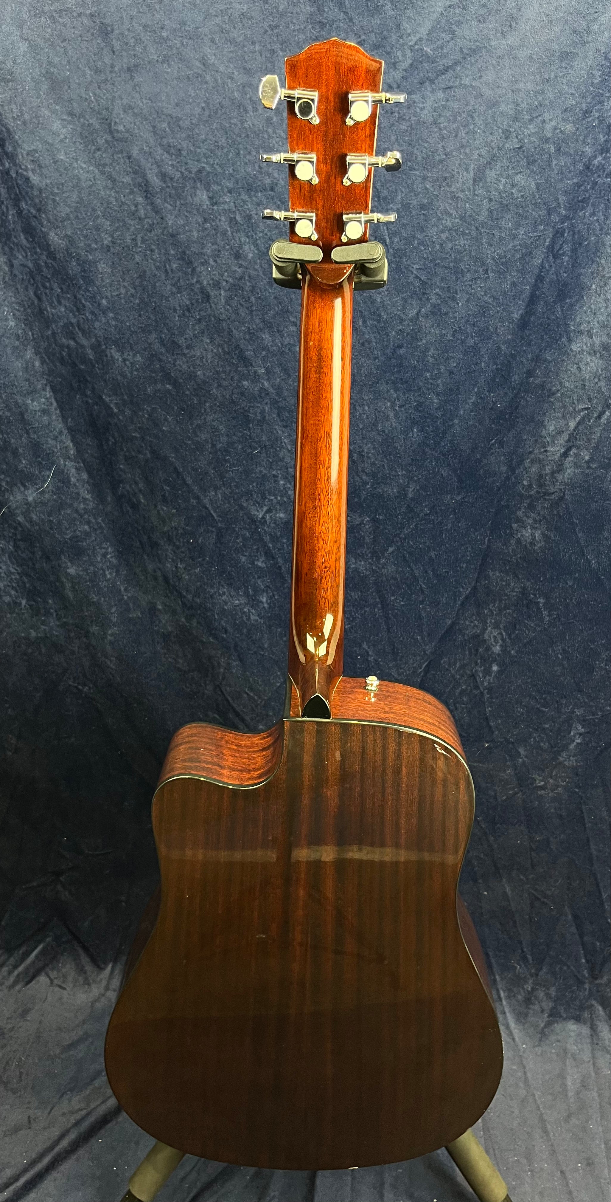 Glossy dark wood acoustic guitar viewed from the back, showing the neck, headstock with six tuners, and the curved body resting on a stand.