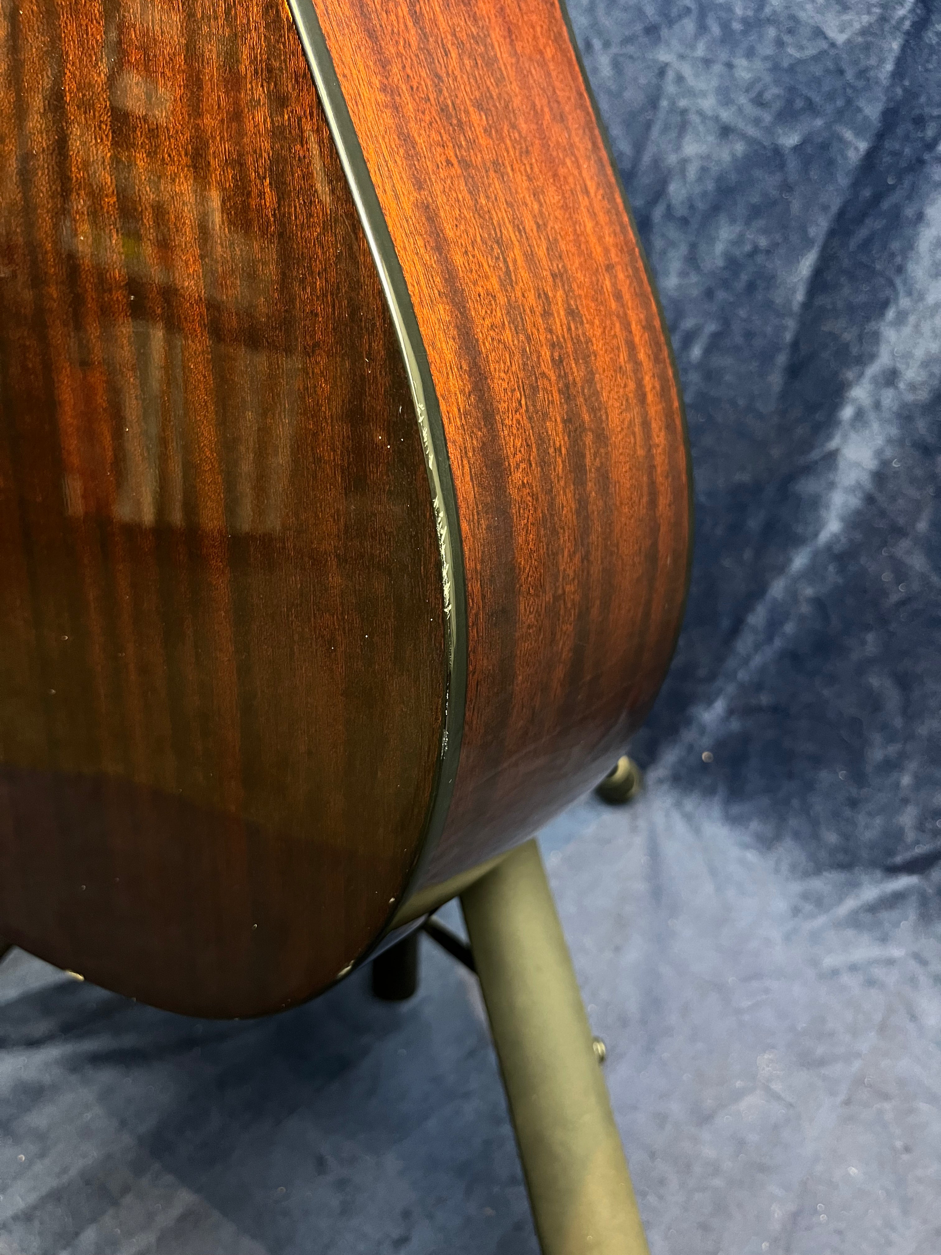Side view of an acoustic guitar showing glossy dark wood with a visible edge scratch near the binding. The guitar rests on a stand.
