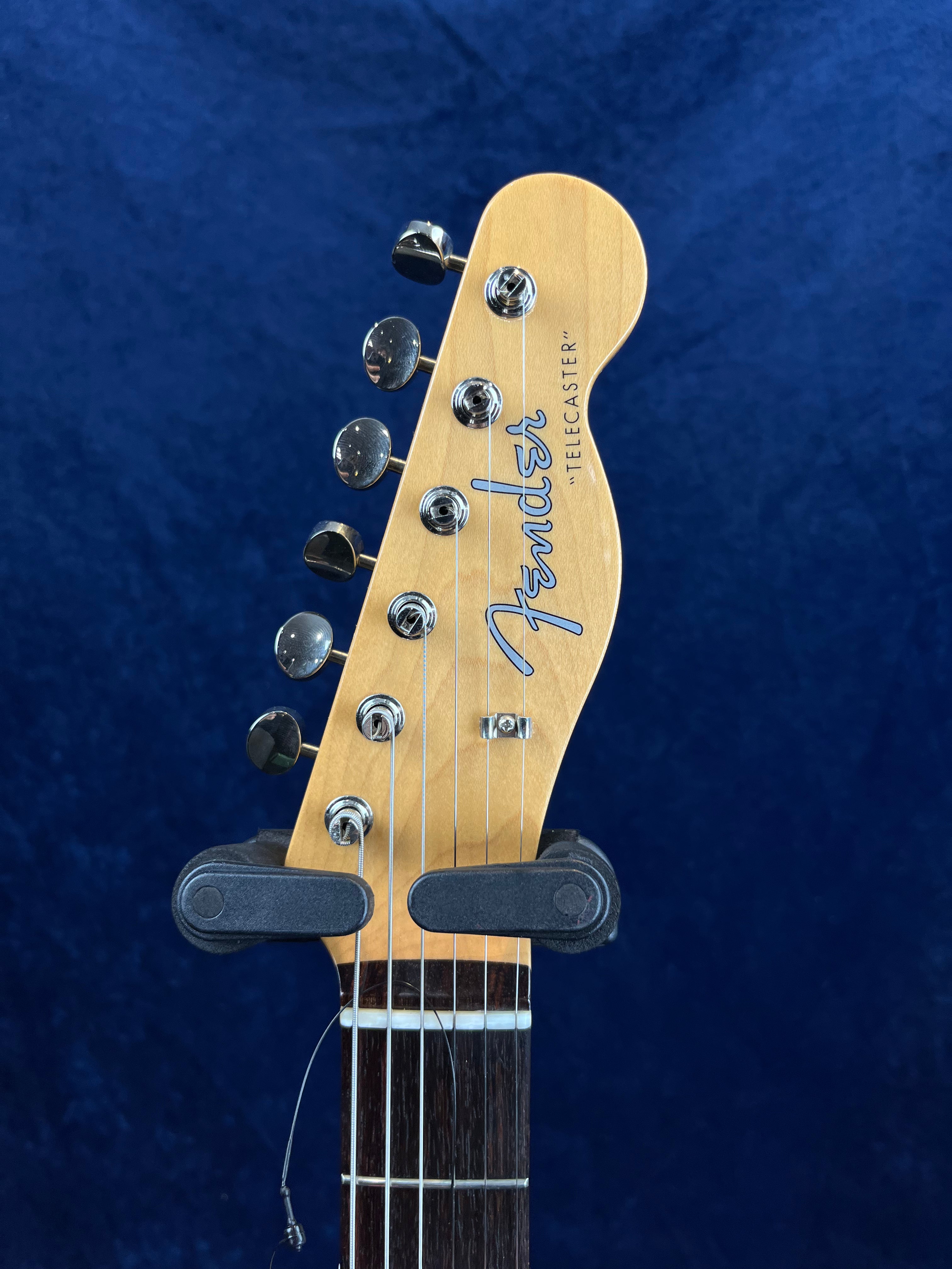 Light wood Fender Telecaster guitar headstock with six tuning pegs, silver "Fender" logo, dark fretboard, against a dark blue background.