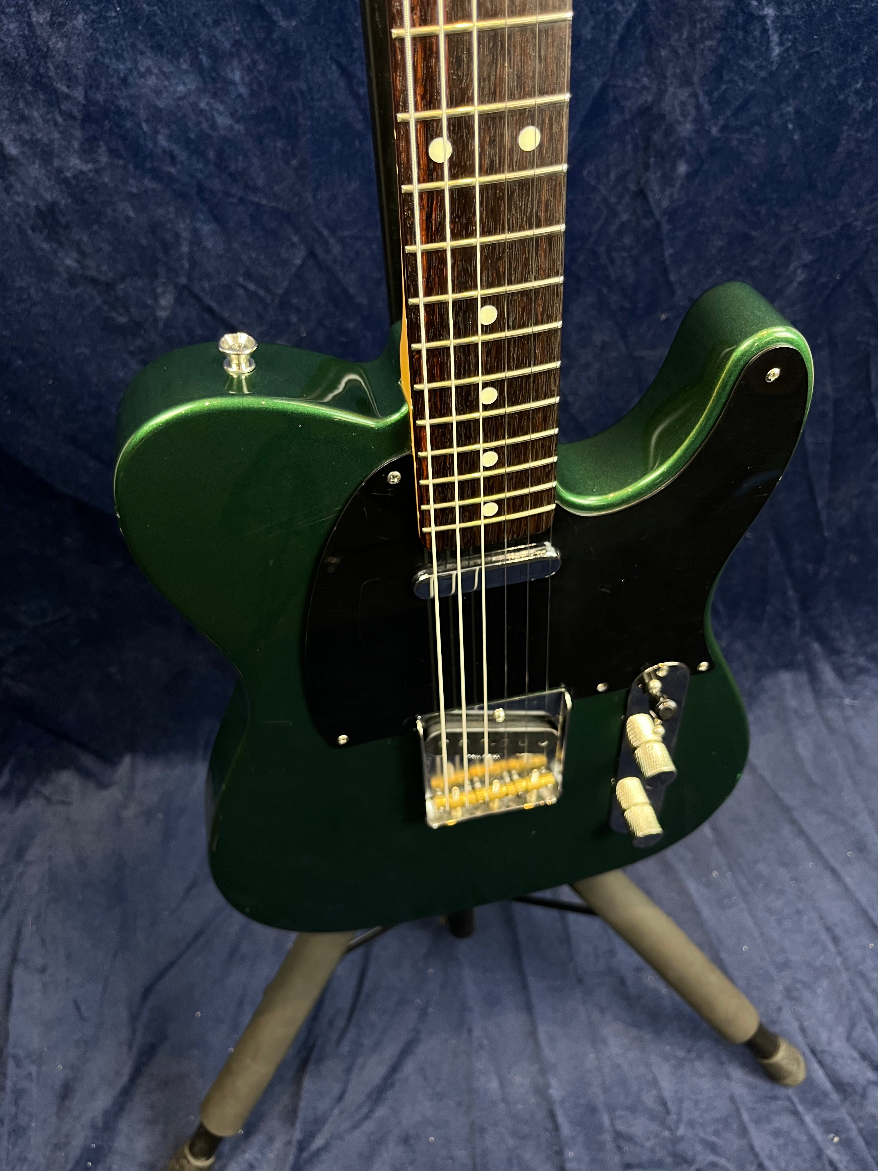 Dark green electric guitar with a black pickguard, two control knobs, and a rosewood fretboard standing on a tripod stand against a dark background.