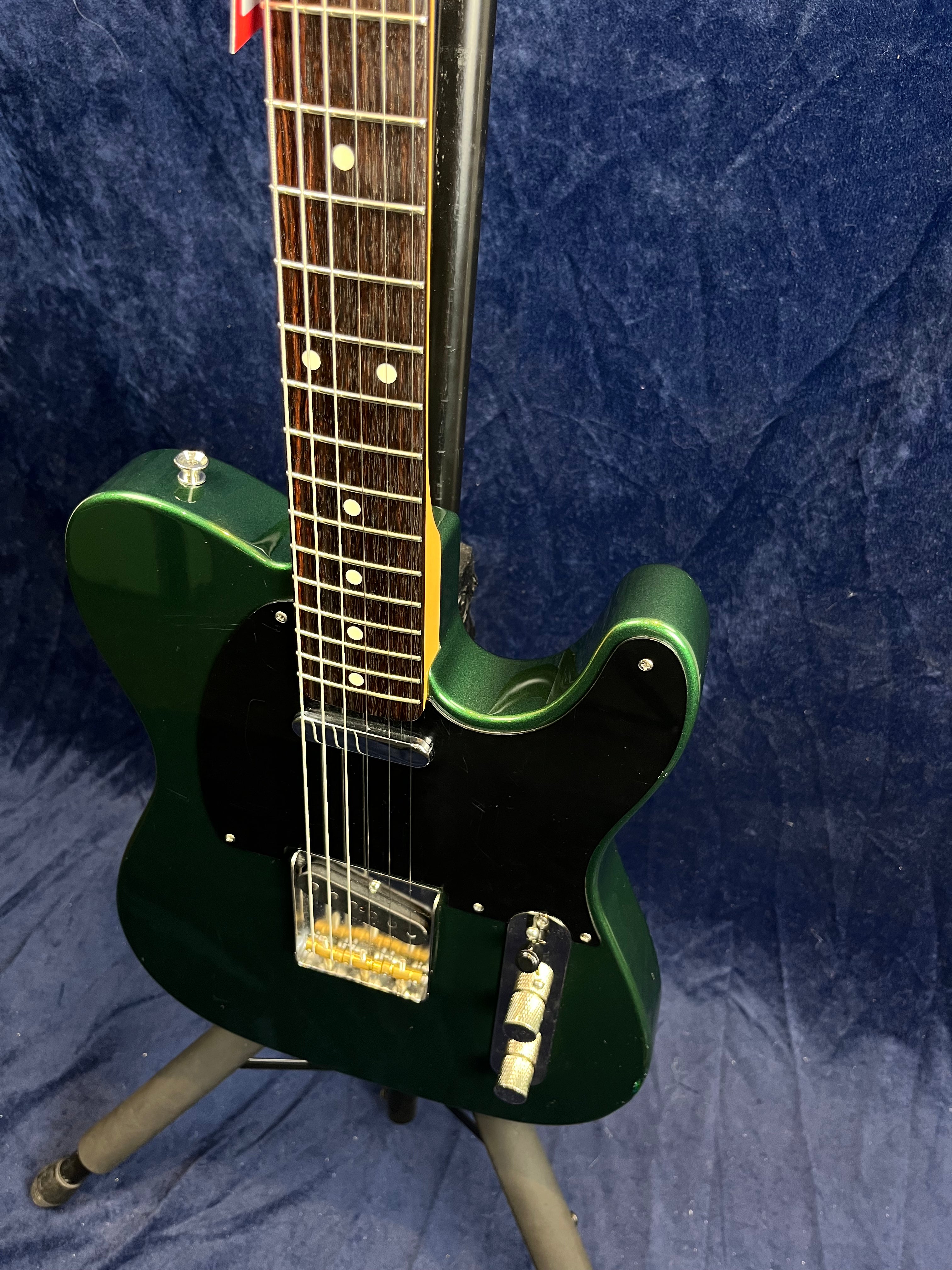 Dark green electric guitar with black pickguard, chrome bridge and control knobs, rosewood fretboard, displayed on a stand against a blue background.