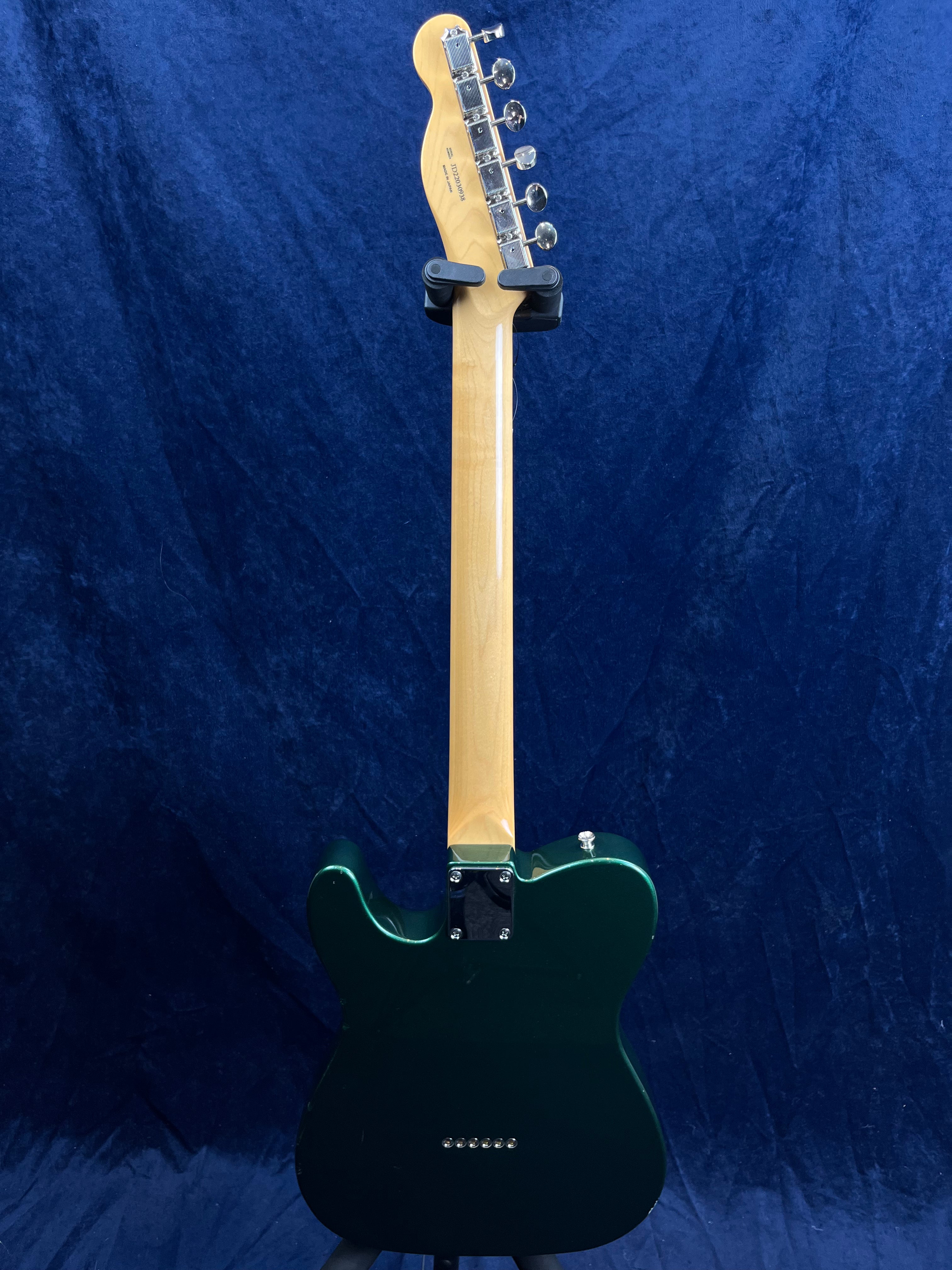 Electric guitar shown from the back with a glossy dark green body, light wood neck, and chrome tuning pegs, set against a textured blue background.