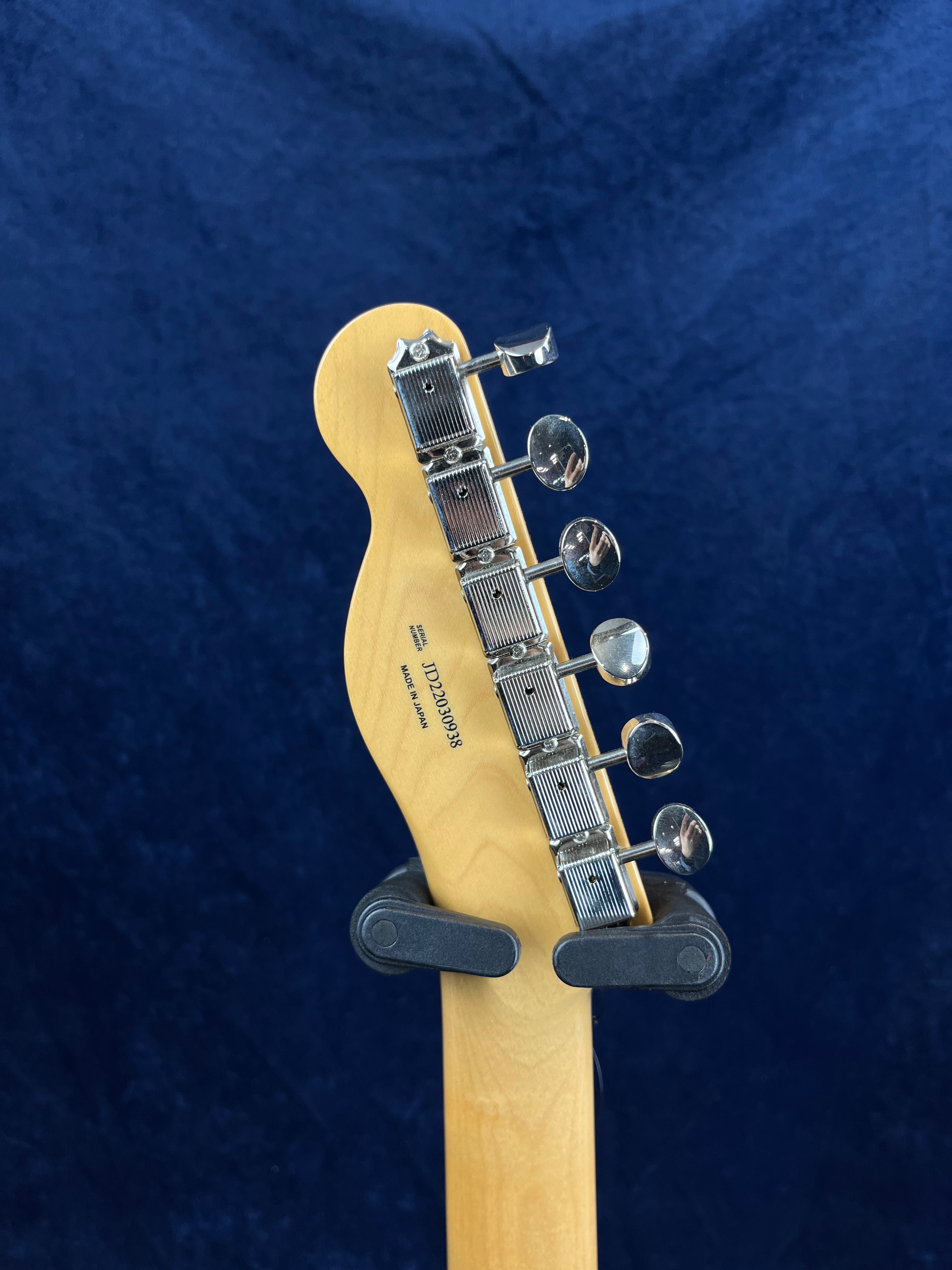 Back of an electric guitar headstock showing six chrome tuning pegs and a visible serial number against a blue background.