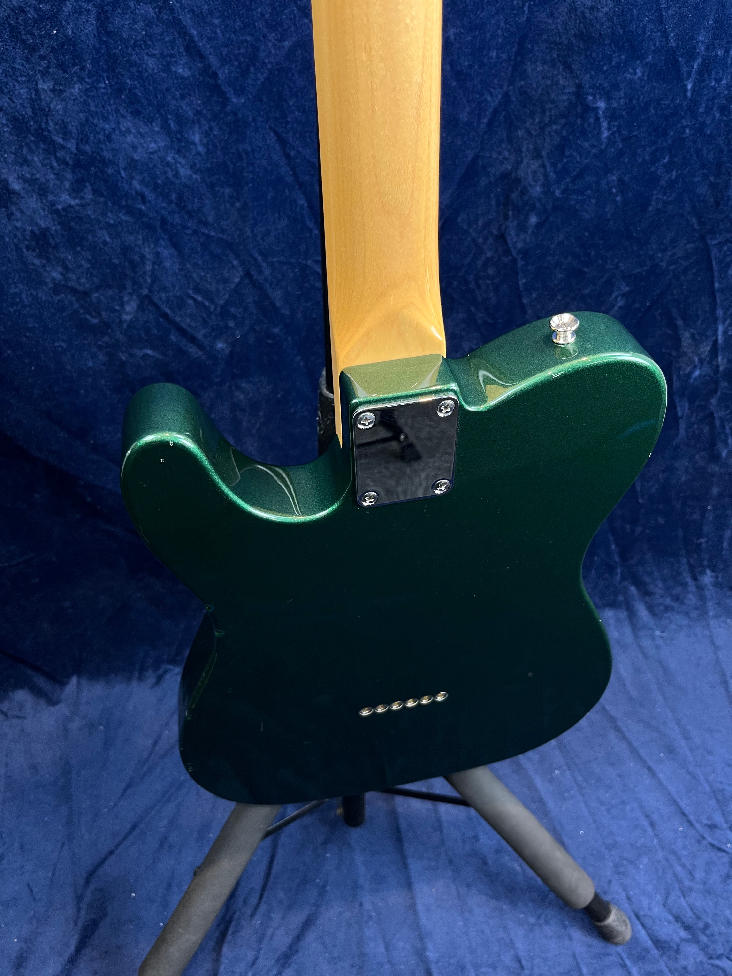Green electric guitar shown from the back, highlighting the neck joint, shiny metallic plate, and string ferrules, standing on a tripod.
