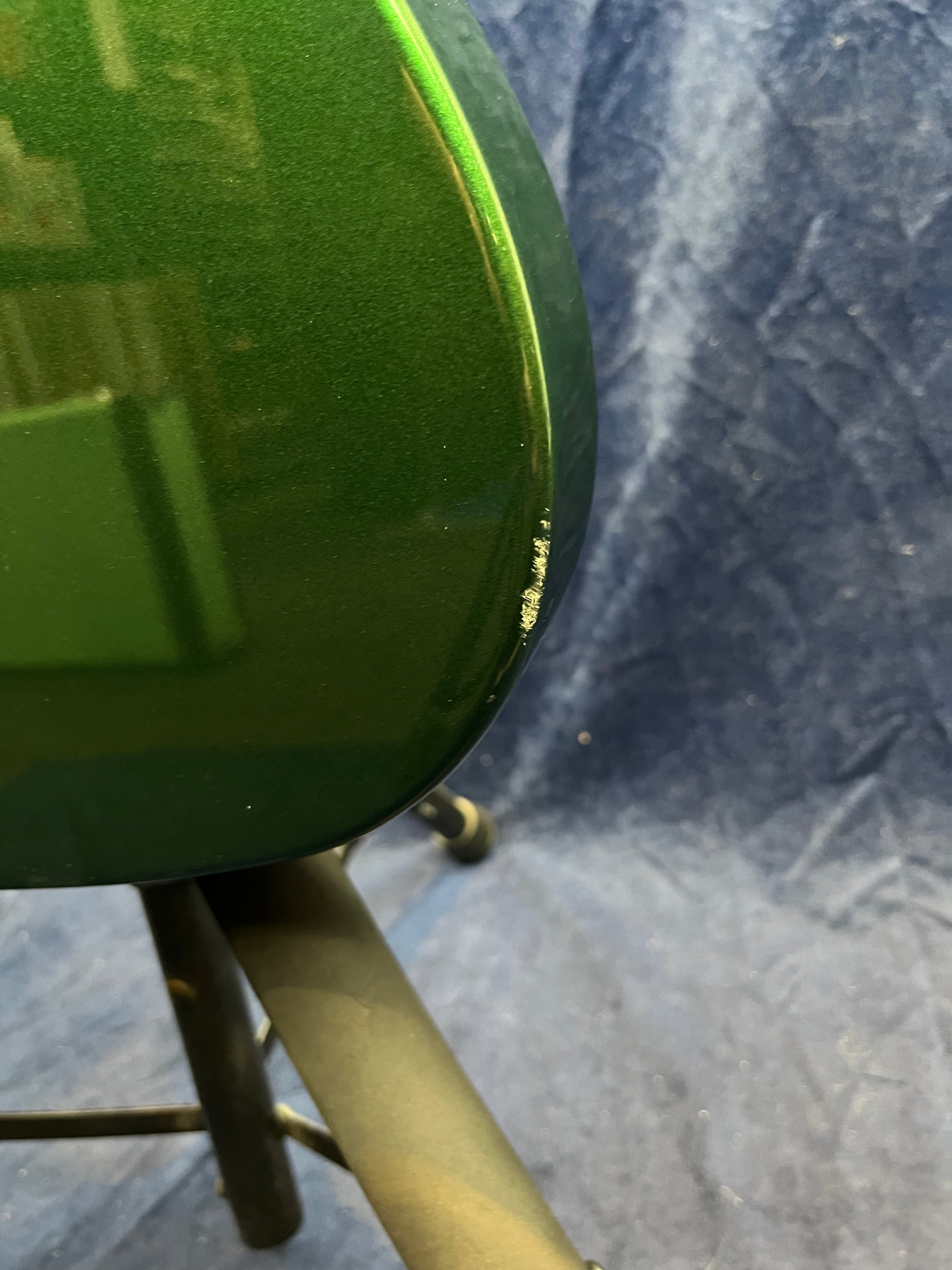 A green electric guitar body with a small area of chipped paint near the bottom edge, shown resting on a stand against a blue background.