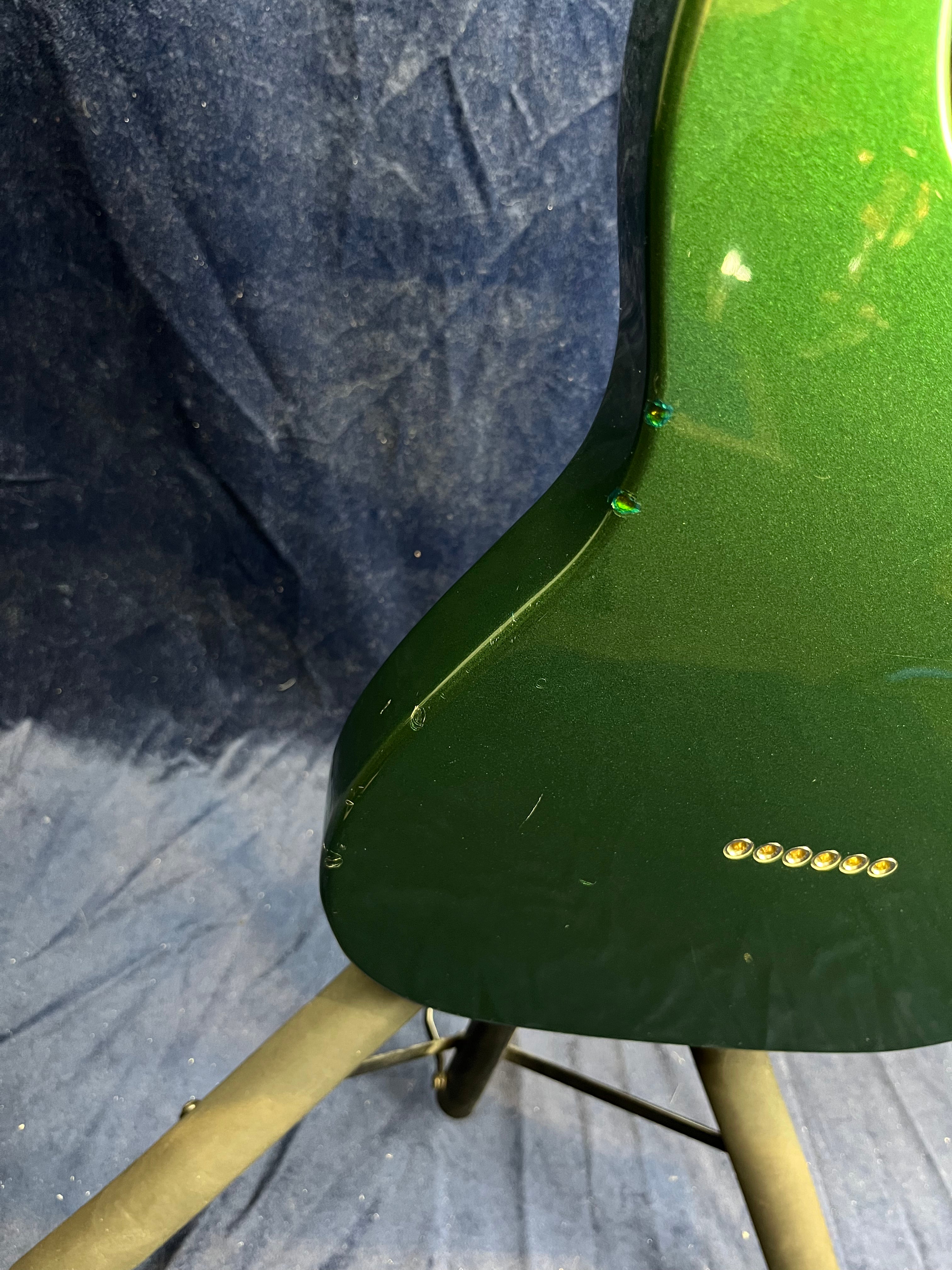 Metallic green electric guitar body with small dings along the edge, six metal string ferrules, and supported by a stand against a dark blue background.