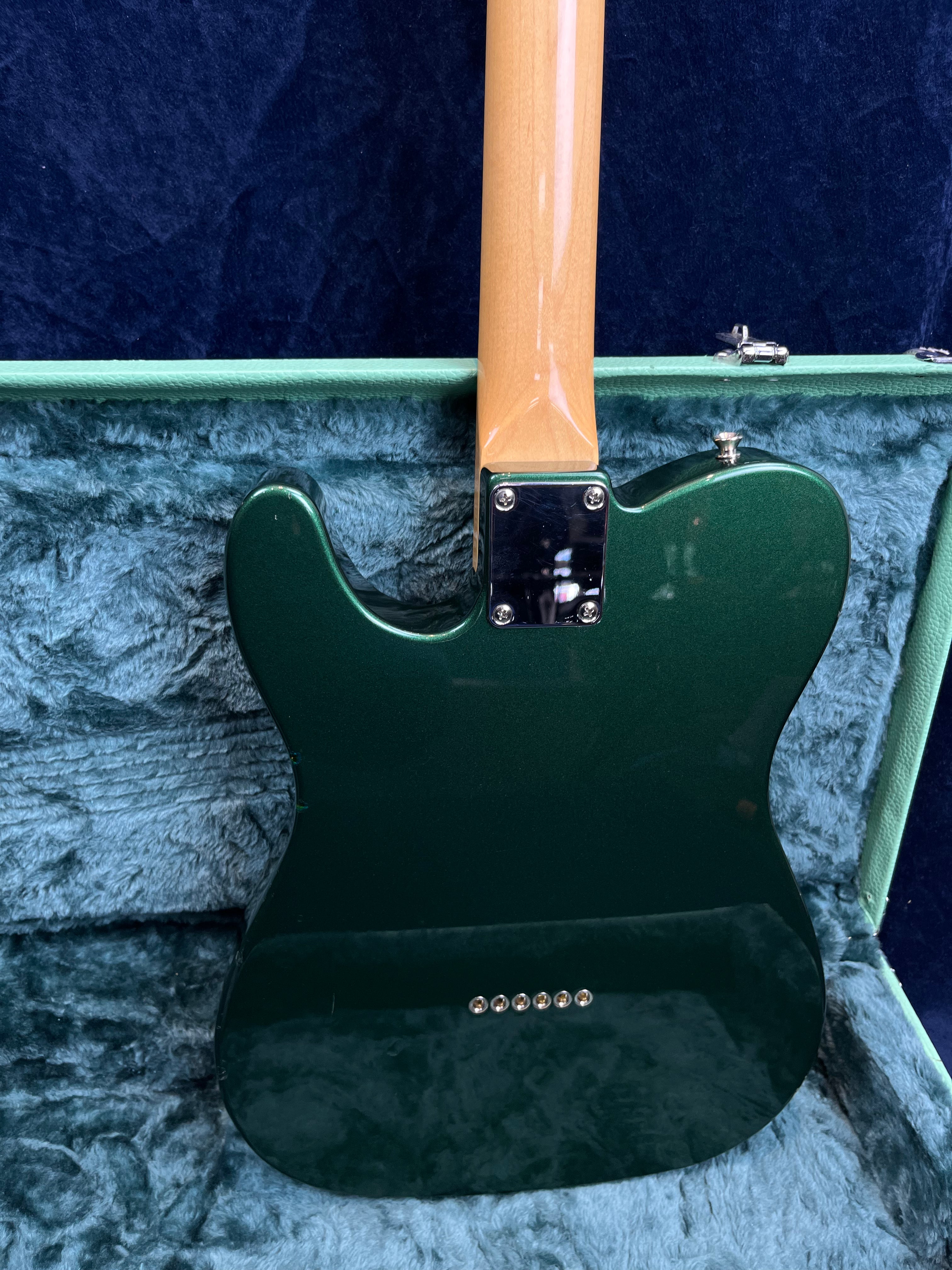 Green electric guitar with a glossy finish, natural wood neck, and six string ferrules on the back, resting inside a blue-lined hard case.