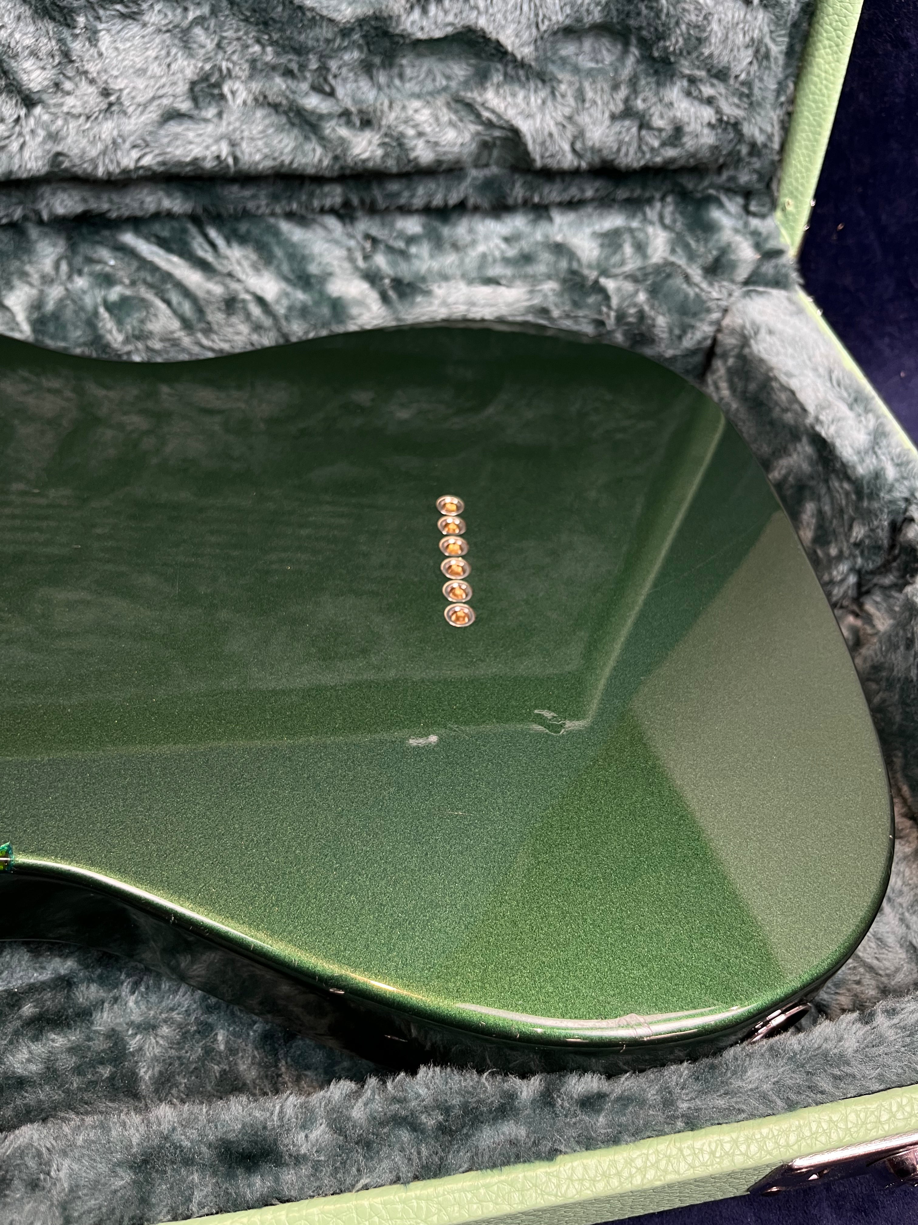 Metallic green electric guitar body with seven gold string ferrules in a row, resting inside a plush, grey-lined hardshell case.
