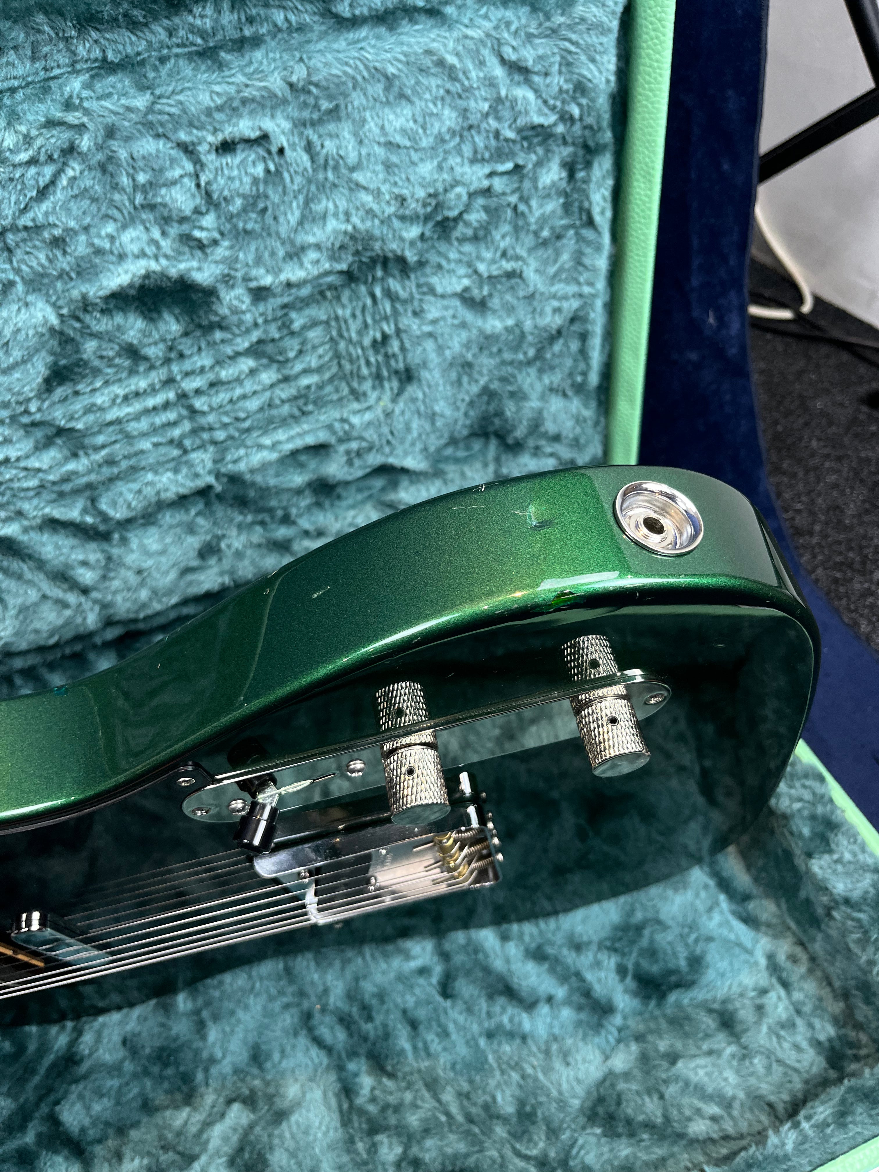 Green electric guitar body close-up showing input jack, two metal control knobs, minor surface scratches, and plush blue-green case interior.