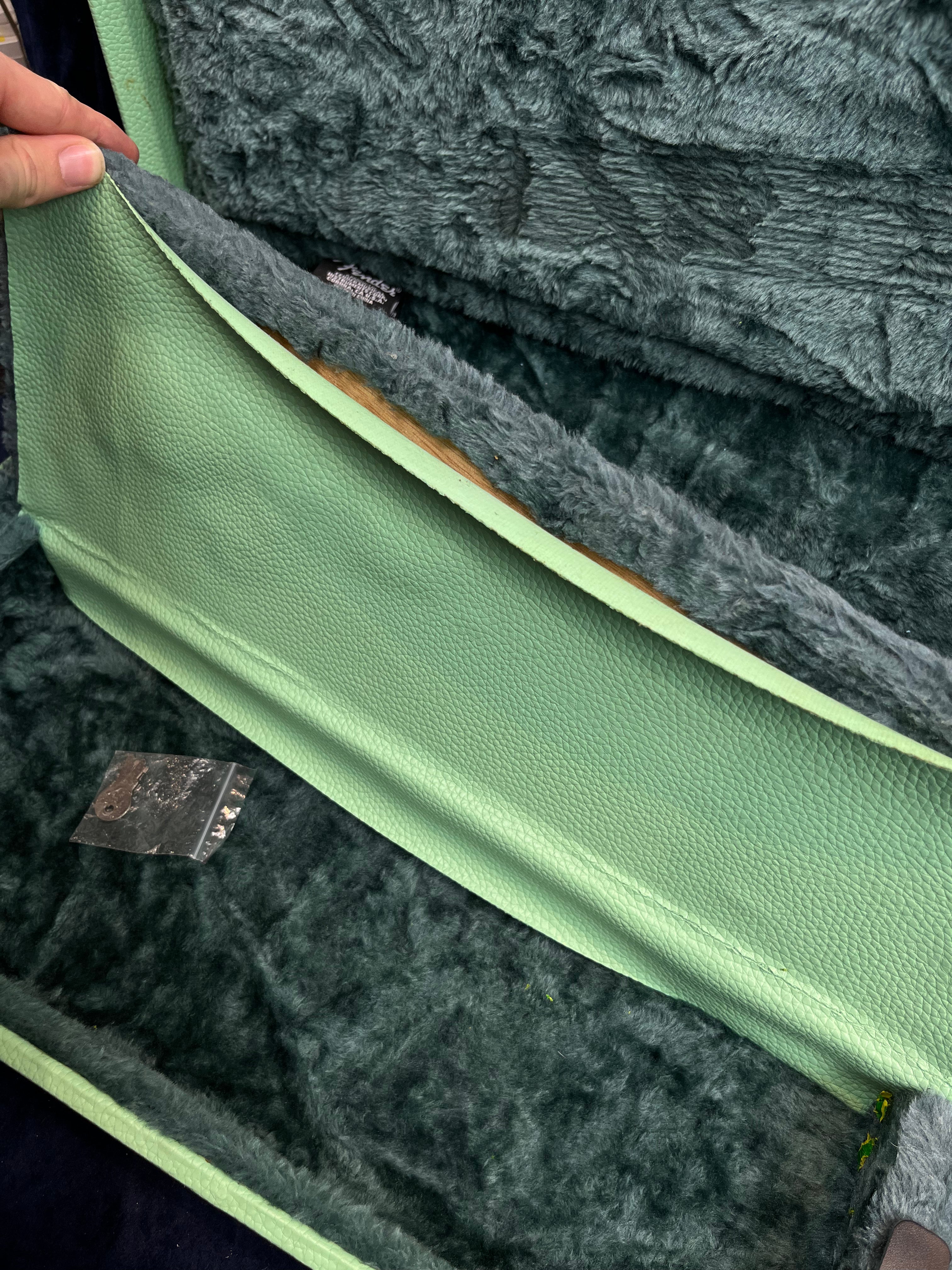 A hand lifts the lining inside a green, plush electric guitar case, revealing a hidden compartment and a small metal bracket inside.