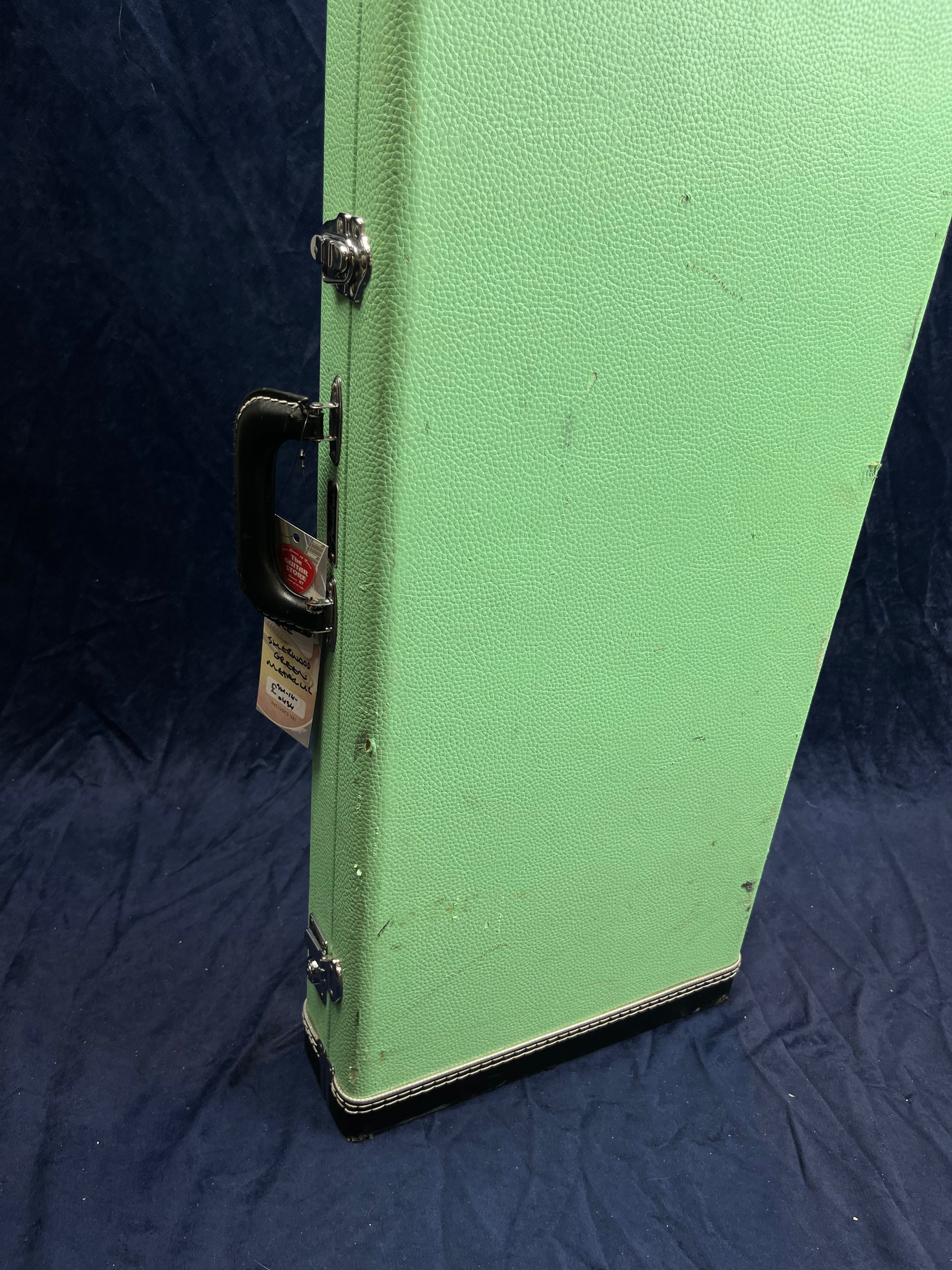 A textured light green electric guitar case stands upright on dark fabric, with metal latches and a black handle attached to a tan tag.