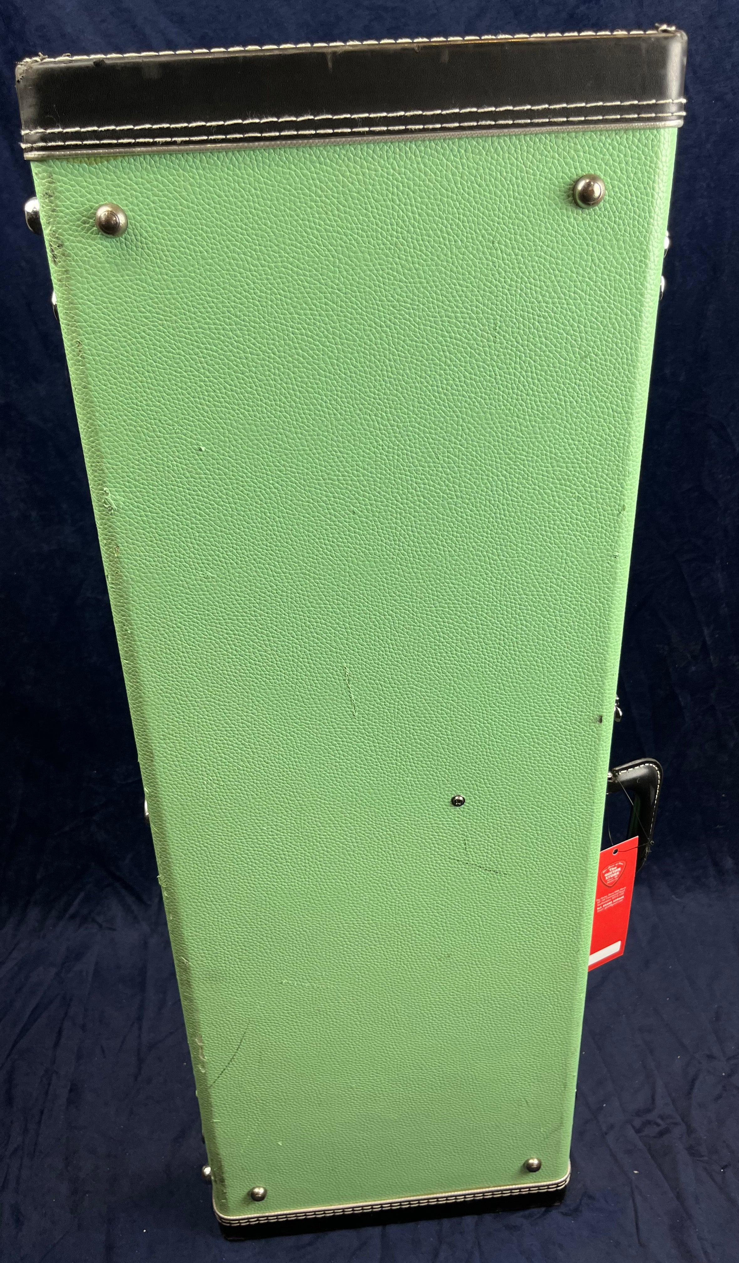 Tall rectangular green electric guitar case with black trim and metal corner protectors, standing upright on a dark background.