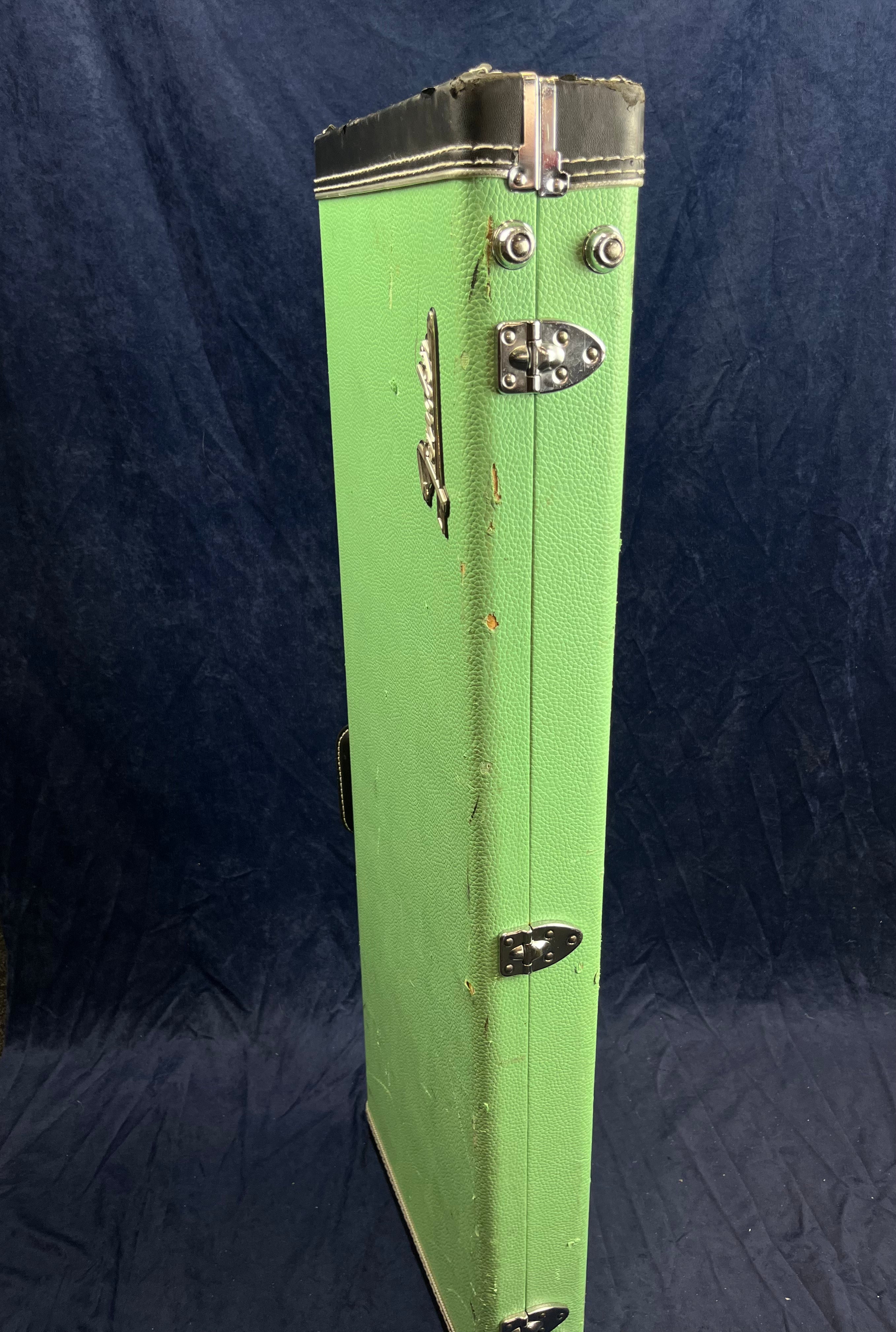 A worn green electric guitar case stands upright with metal latches, black edges, and some scratches and dents visible on its surface.