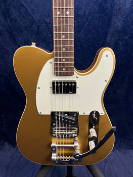 Gold electric guitar with white pickguard, two pickups, Bigsby-style tremolo, chrome knobs, and rosewood fingerboard against a dark background.