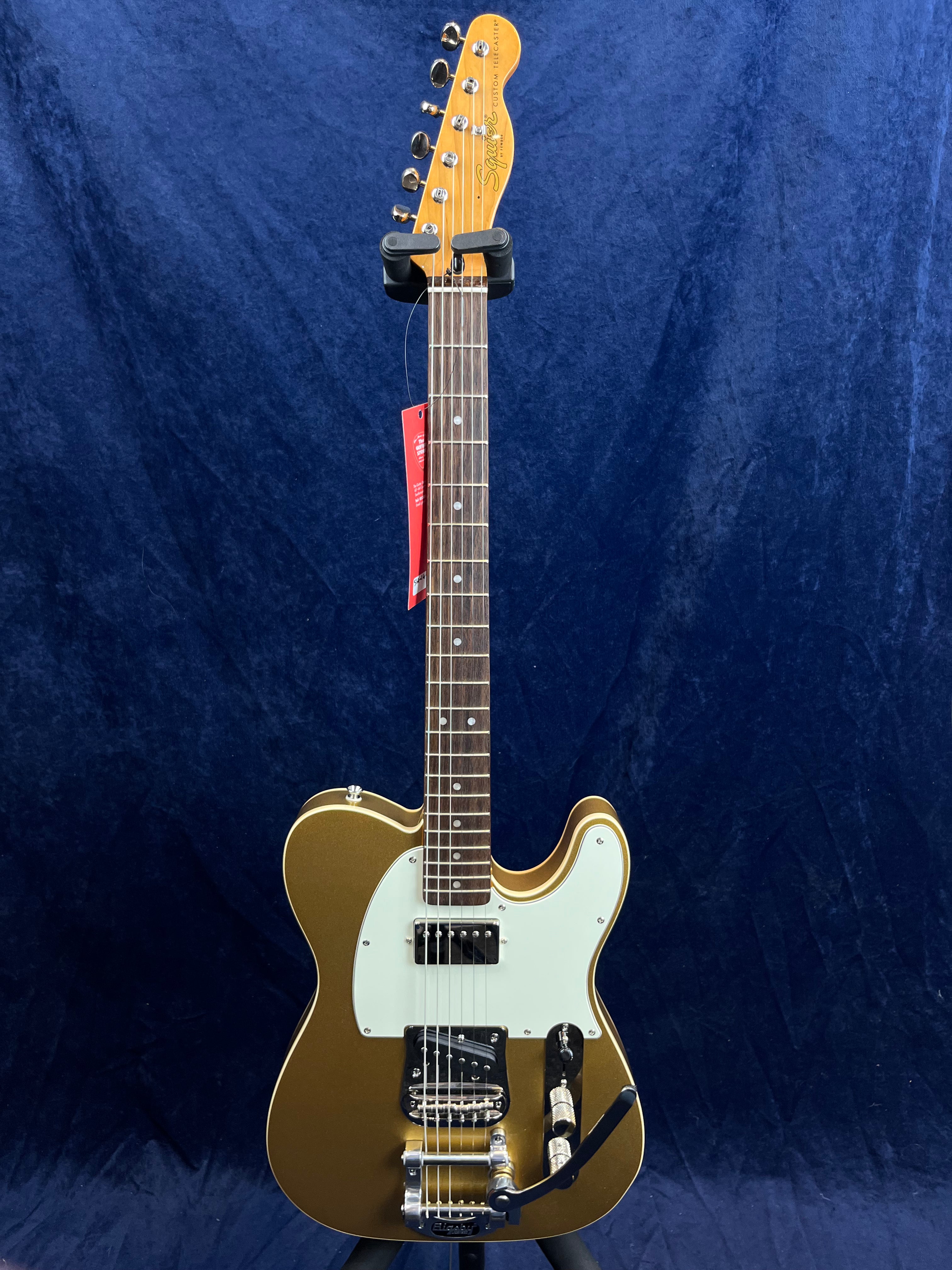 Gold Squier Telecaster-style electric guitar with white pickguard, two pickups, chrome hardware, tremolo arm, and rosewood fretboard against dark blue.