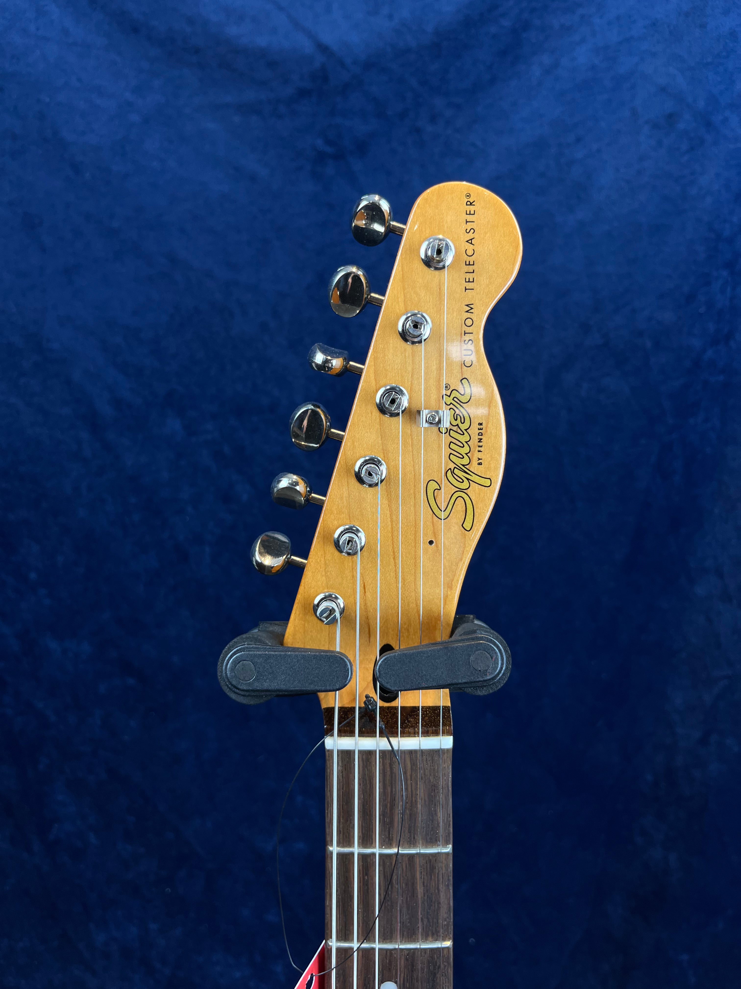 Headstock of a Squier Custom Telecaster electric guitar with chrome tuning pegs, visible strings, and a dark wood fretboard against a blue background.
