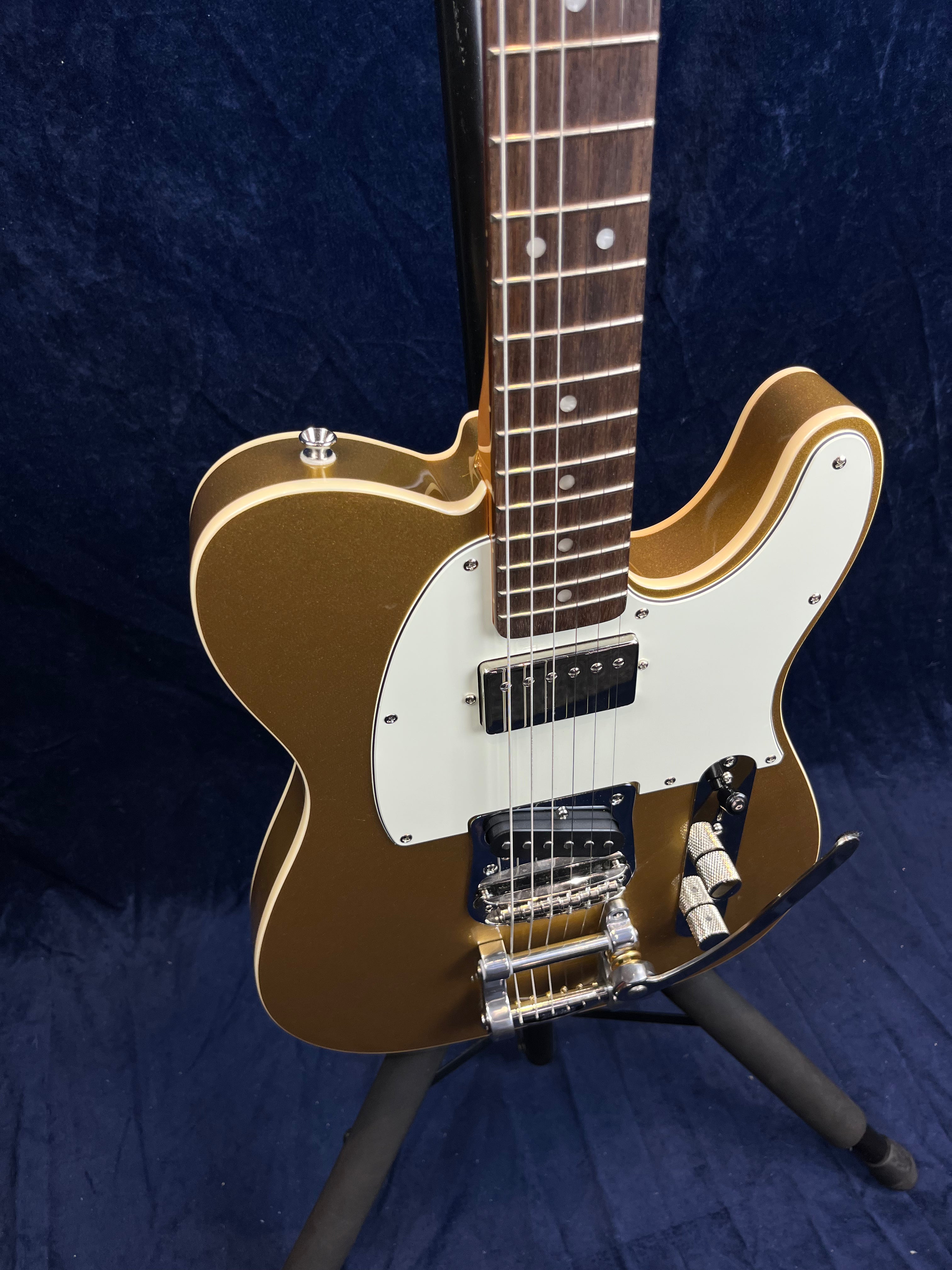 Gold electric guitar with white pickguard, two pickups, and a silver tremolo arm, shown on a stand against a dark blue background.