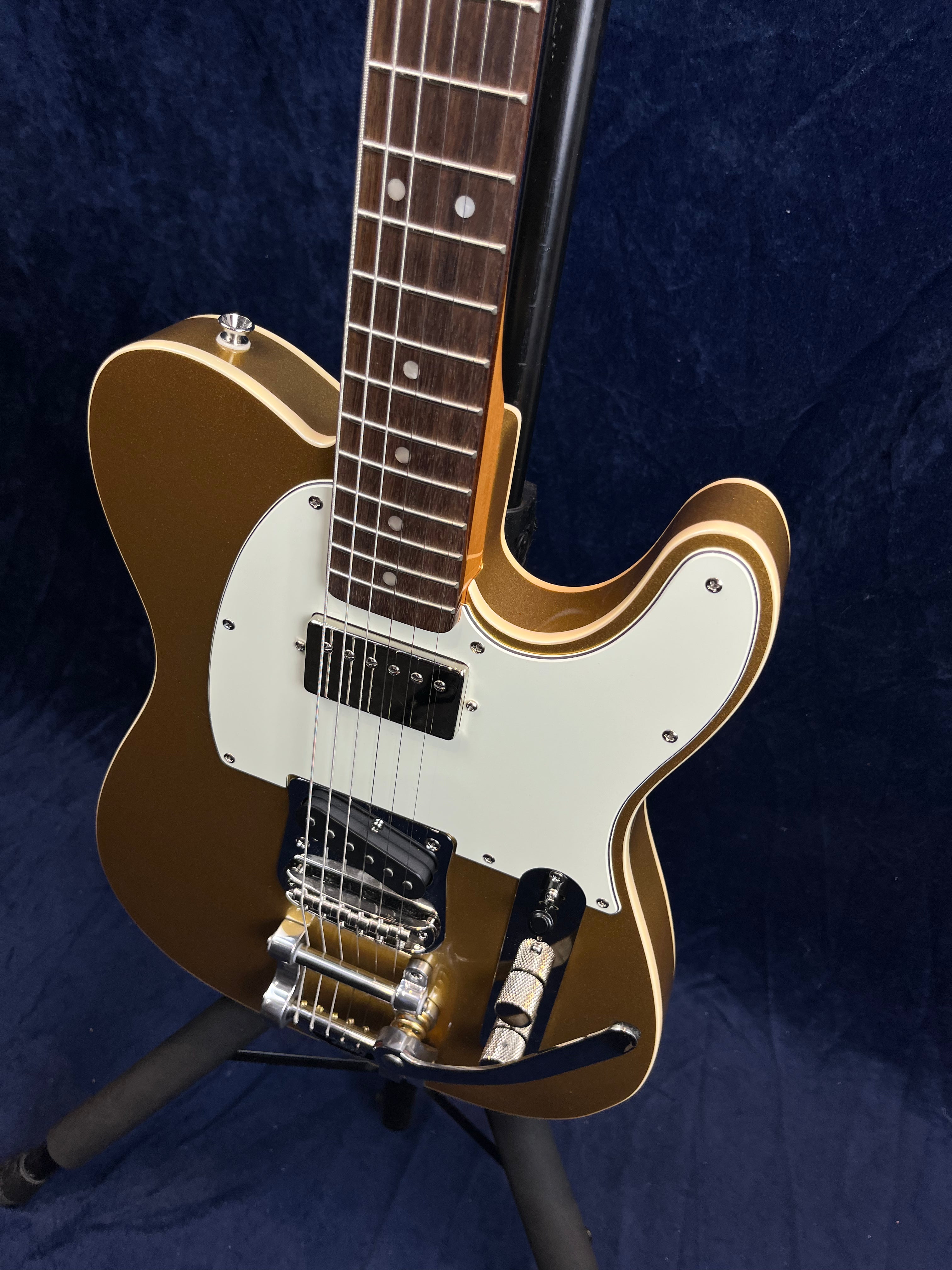 Gold electric guitar with a white pickguard, chrome hardware, a tremolo bridge, and a rosewood fingerboard on a stand against a dark background.