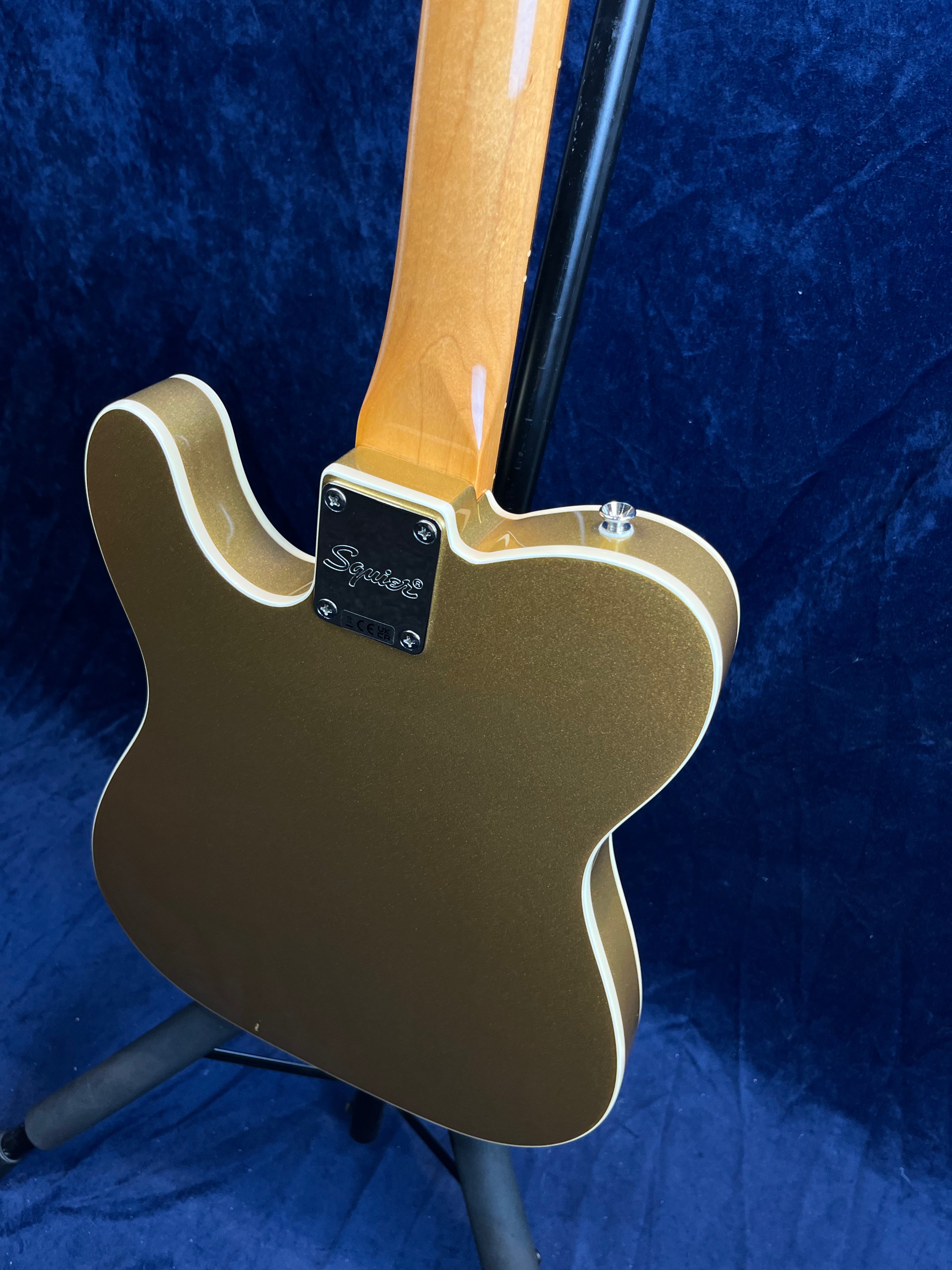 Gold Squier electric guitar shown from the back, with a glossy finish, white binding, and a natural wood neck against a dark blue background.