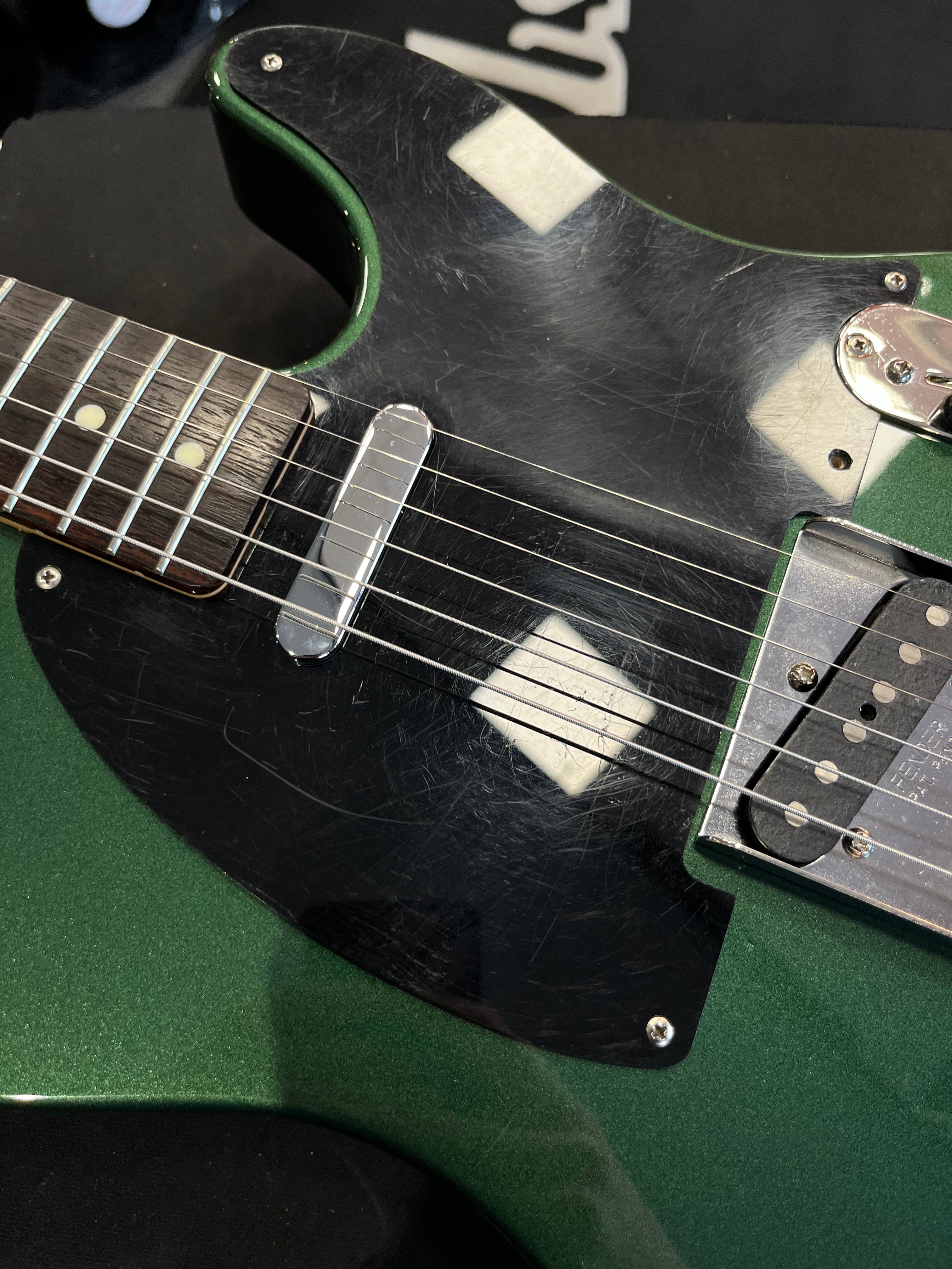 Metallic green electric guitar body with a black, heavily scratched pickguard, chrome pickup covers, and visible white reflections.