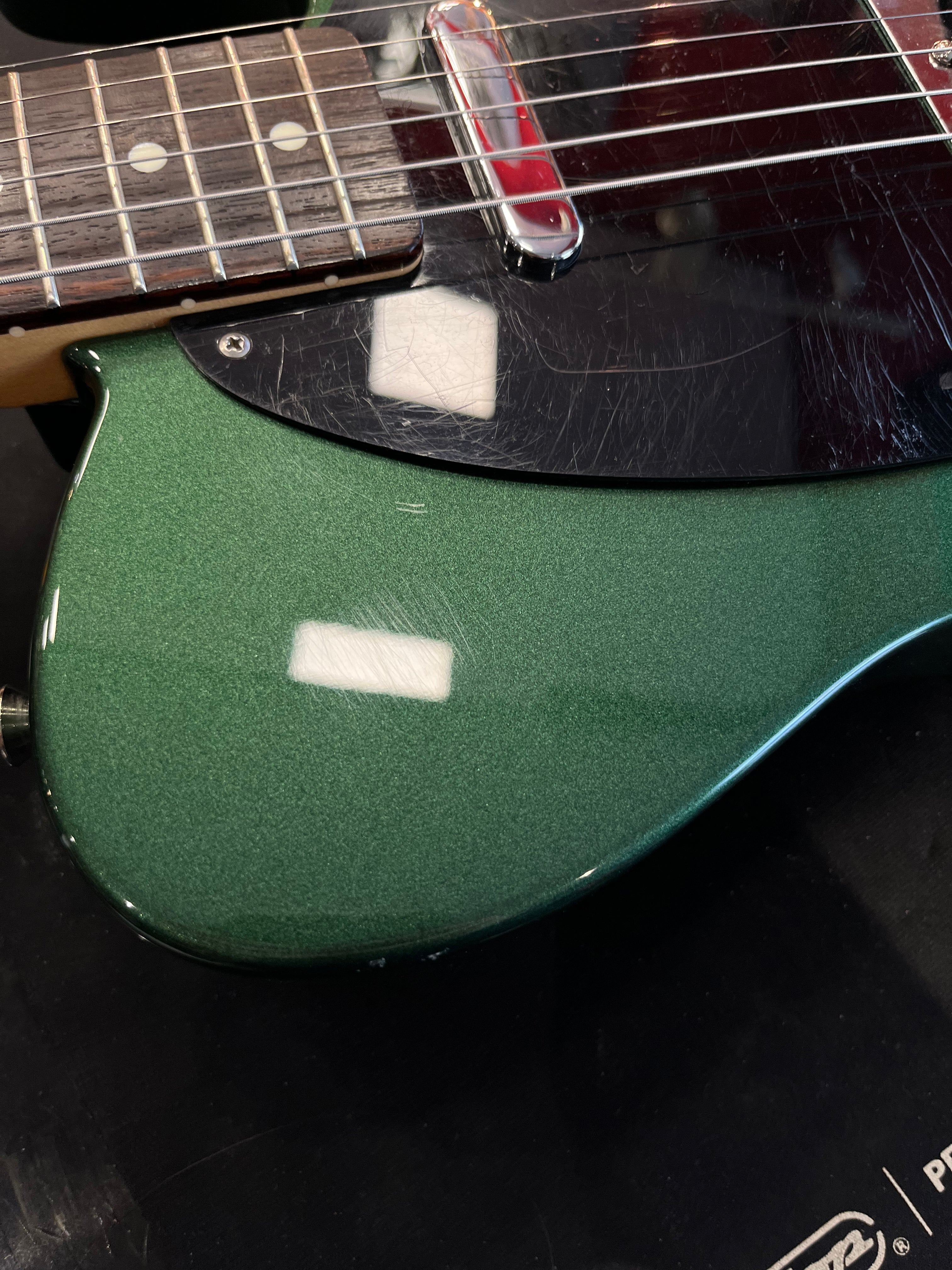 Green electric guitar body with a black pickguard showing many scratches, metal pickup, and part of a rosewood fretboard with dot inlays.