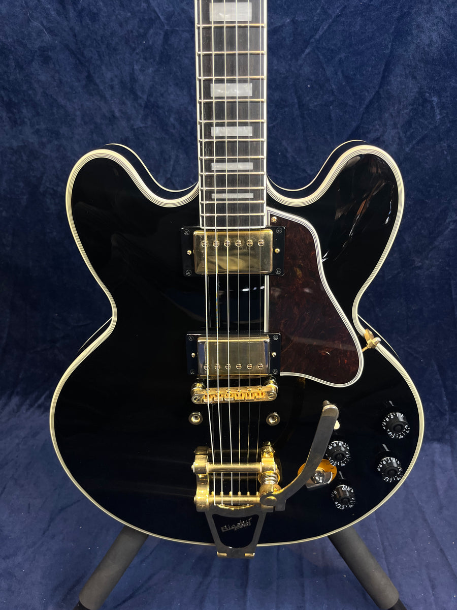Black semi-hollow electric guitar with gold hardware, two humbucker pickups, four control knobs, and a Bigsby vibrato tailpiece.