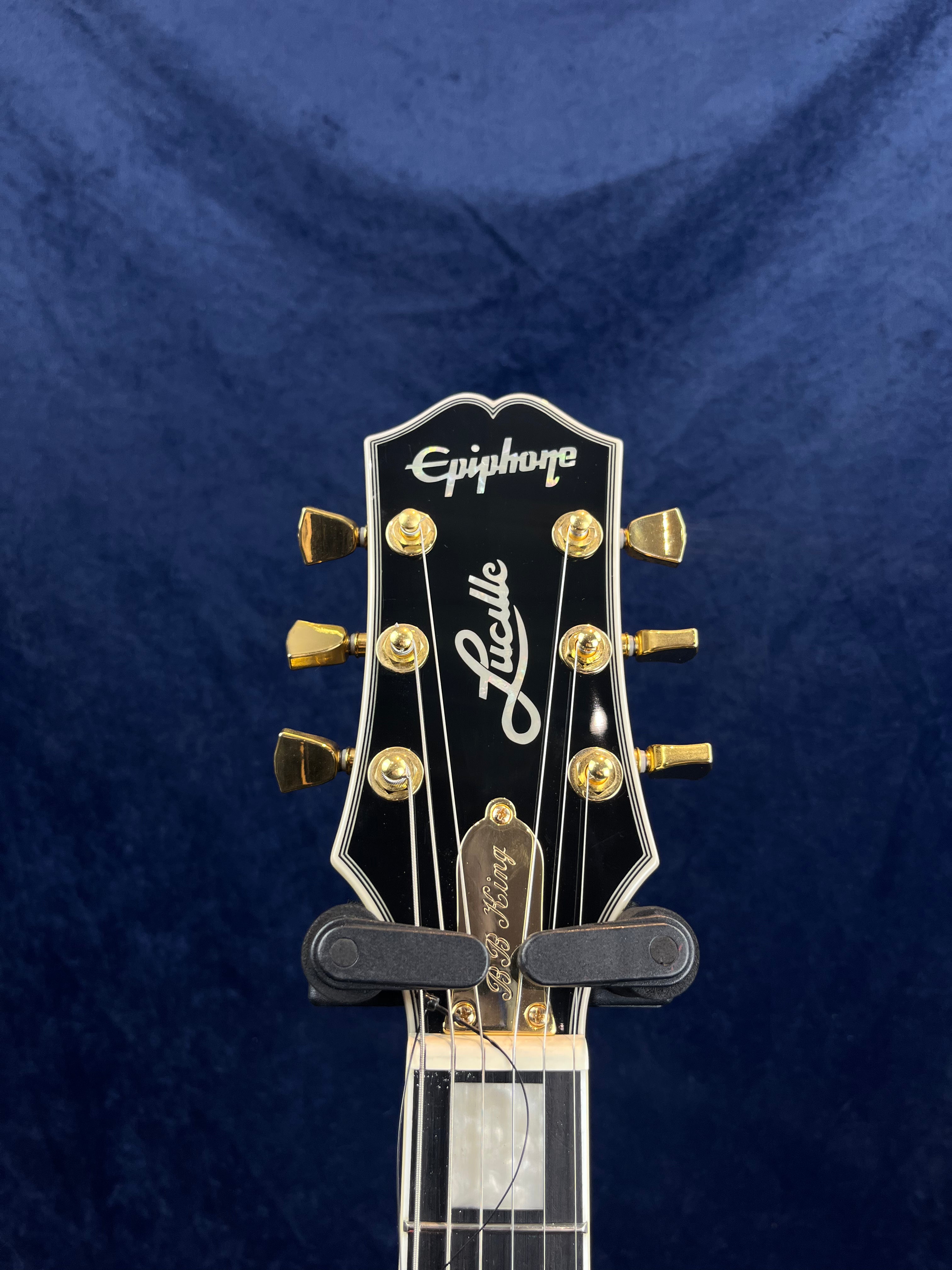 Epiphone Lucille electric guitar headstock with gold tuning pegs, black finish, and “Lucille” script on the front against a dark blue background.