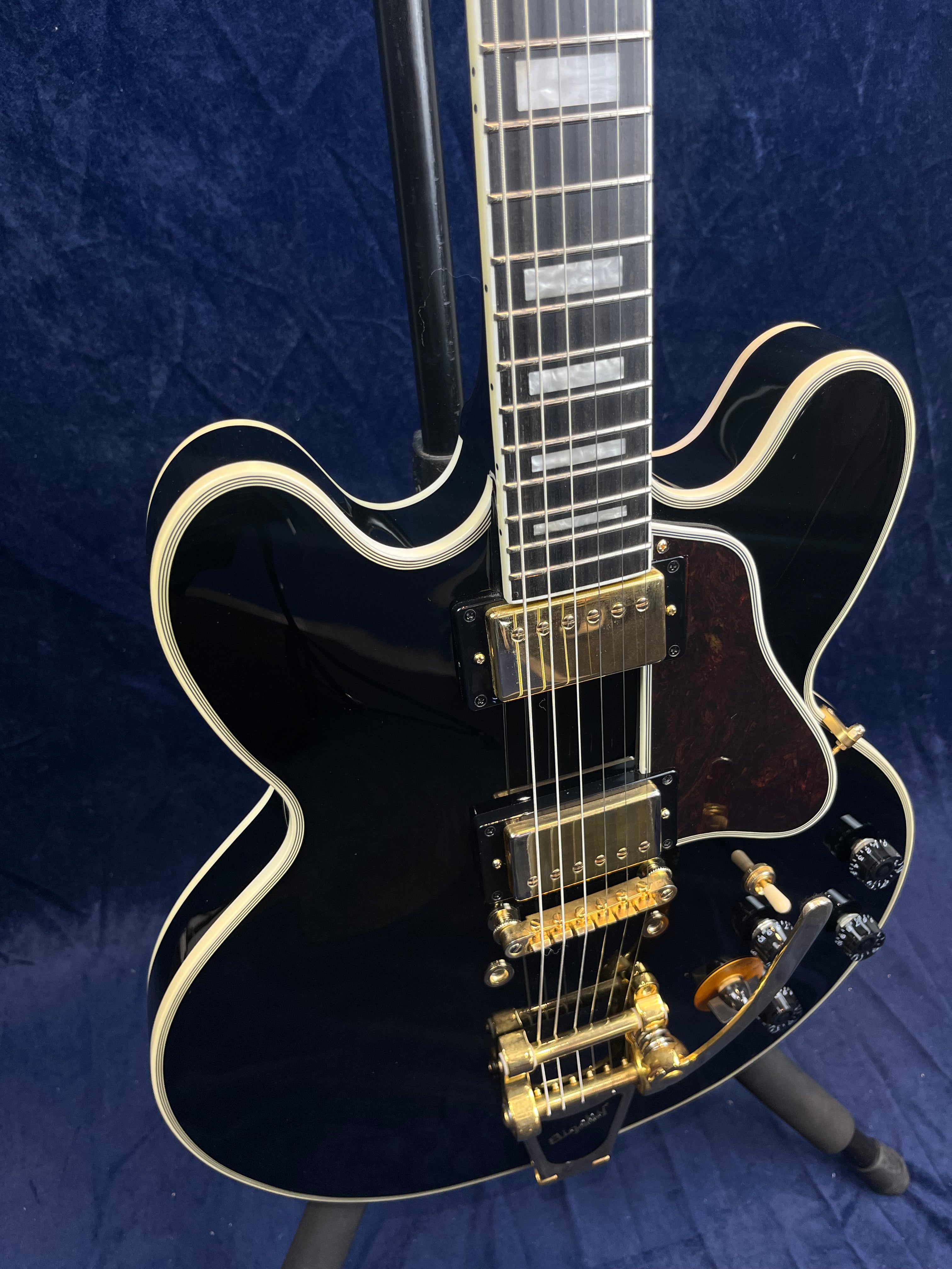 Black electric guitar with gold hardware, cream binding, two humbucker pickups, Bigsby-style tremolo, and rectangular fretboard inlays.