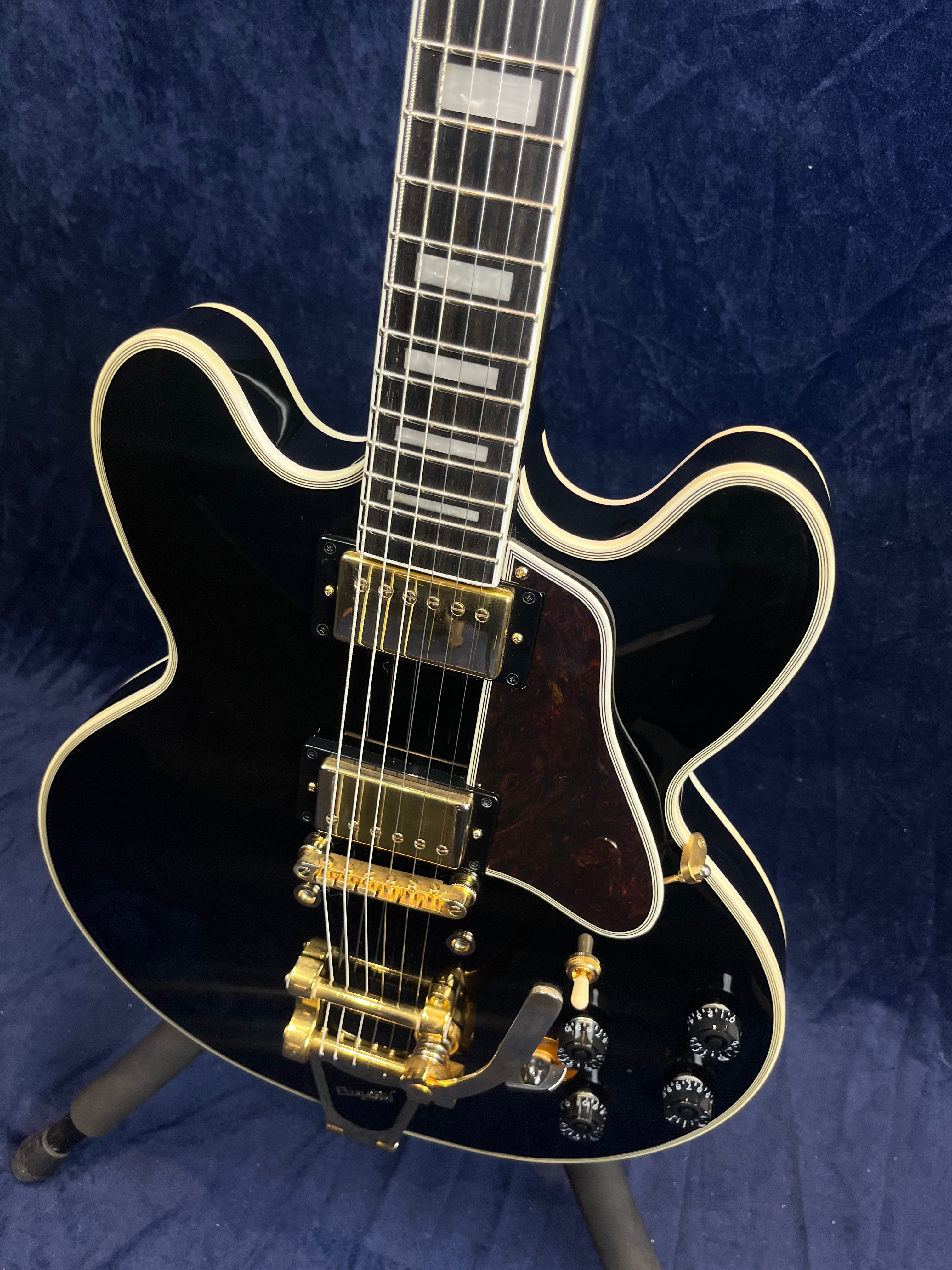 Black semi-hollow electric guitar with gold hardware, block inlays on the neck, multiple control knobs, and a tremolo tailpiece on a stand.