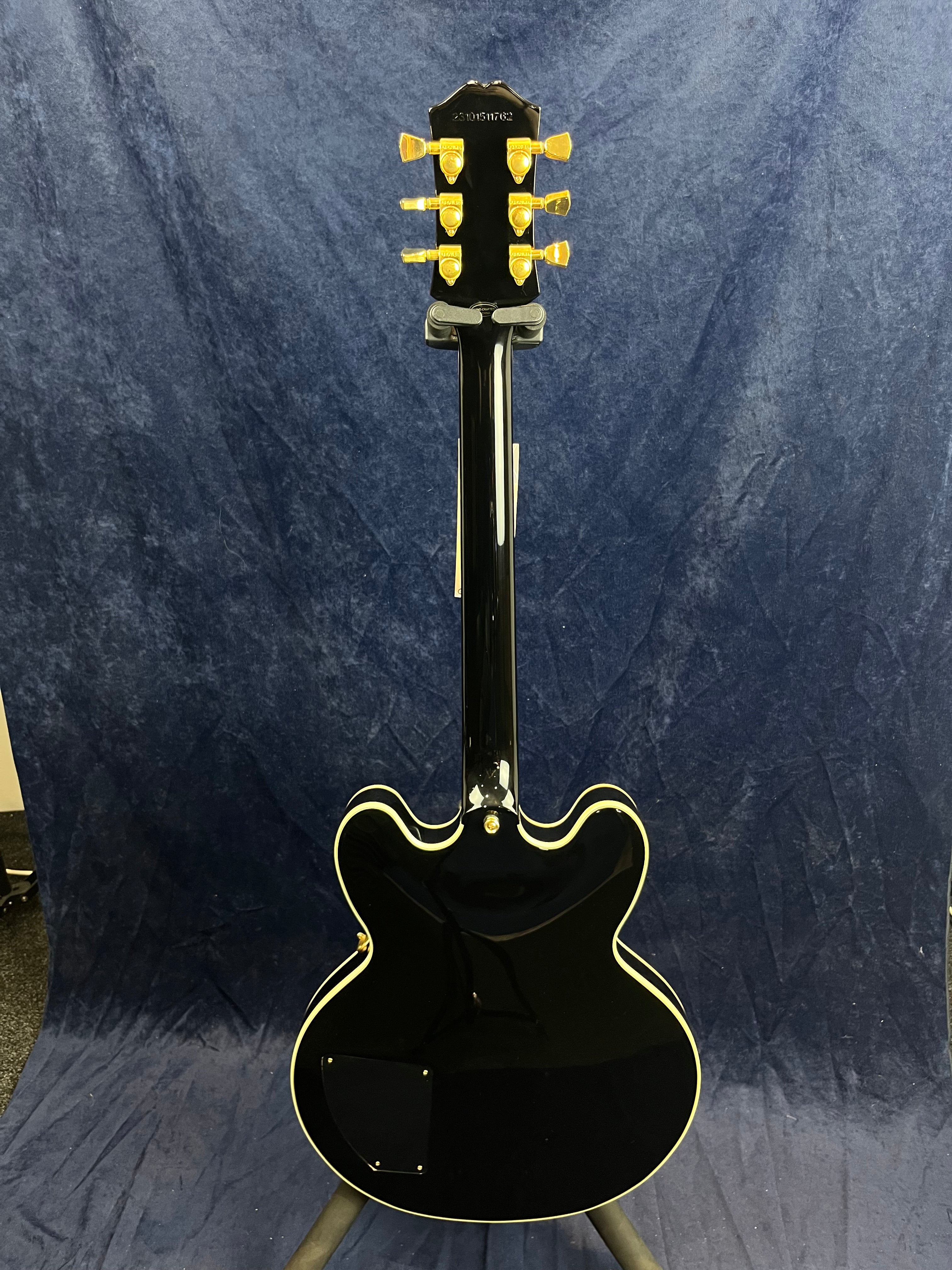 Glossy black electric guitar seen from the back, double-cutaway body, cream binding, gold tuning pegs, and set against a dark blue background.