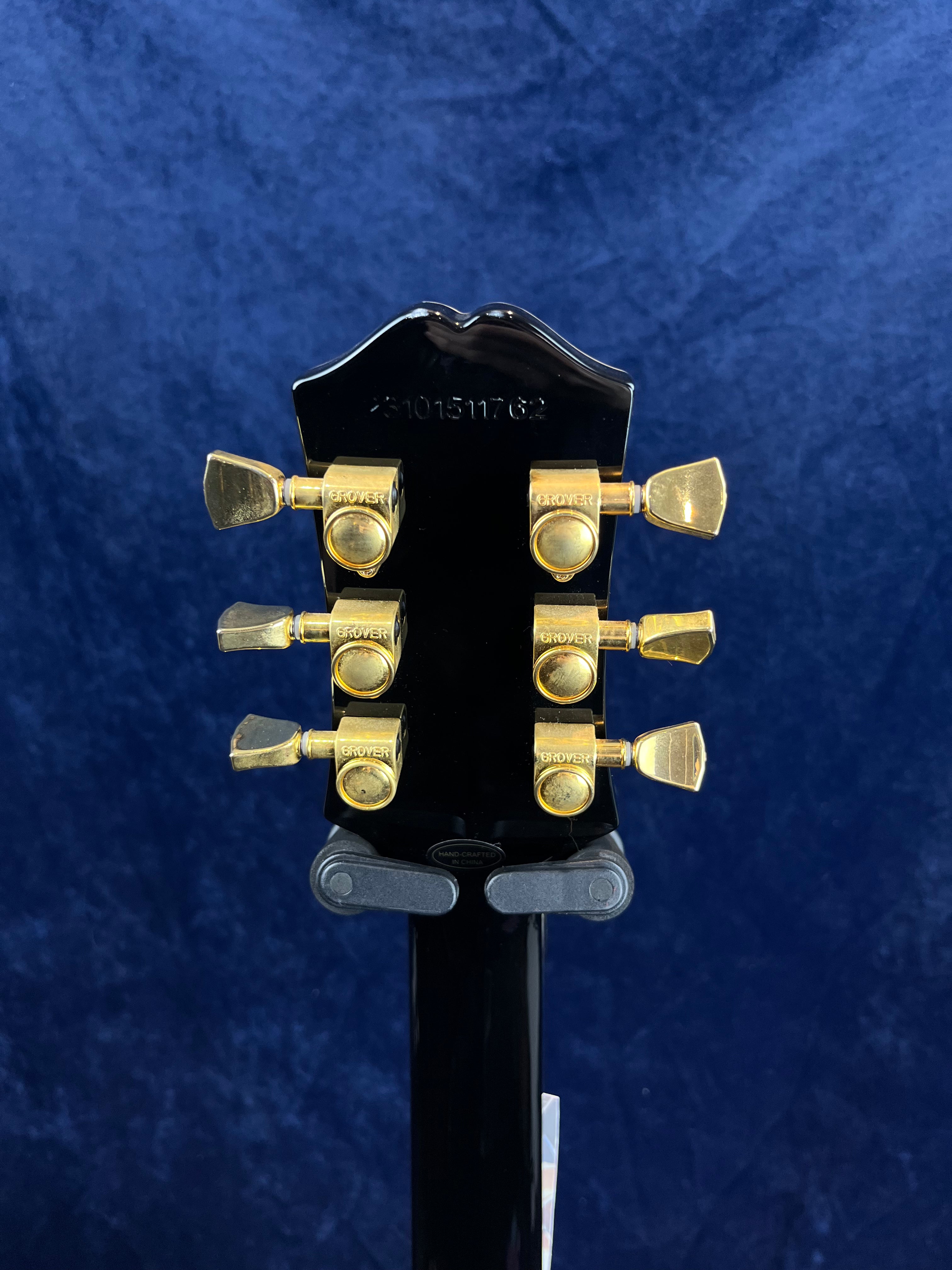 Back of a black electric guitar headstock with six gold Grover tuning pegs against a dark blue background.