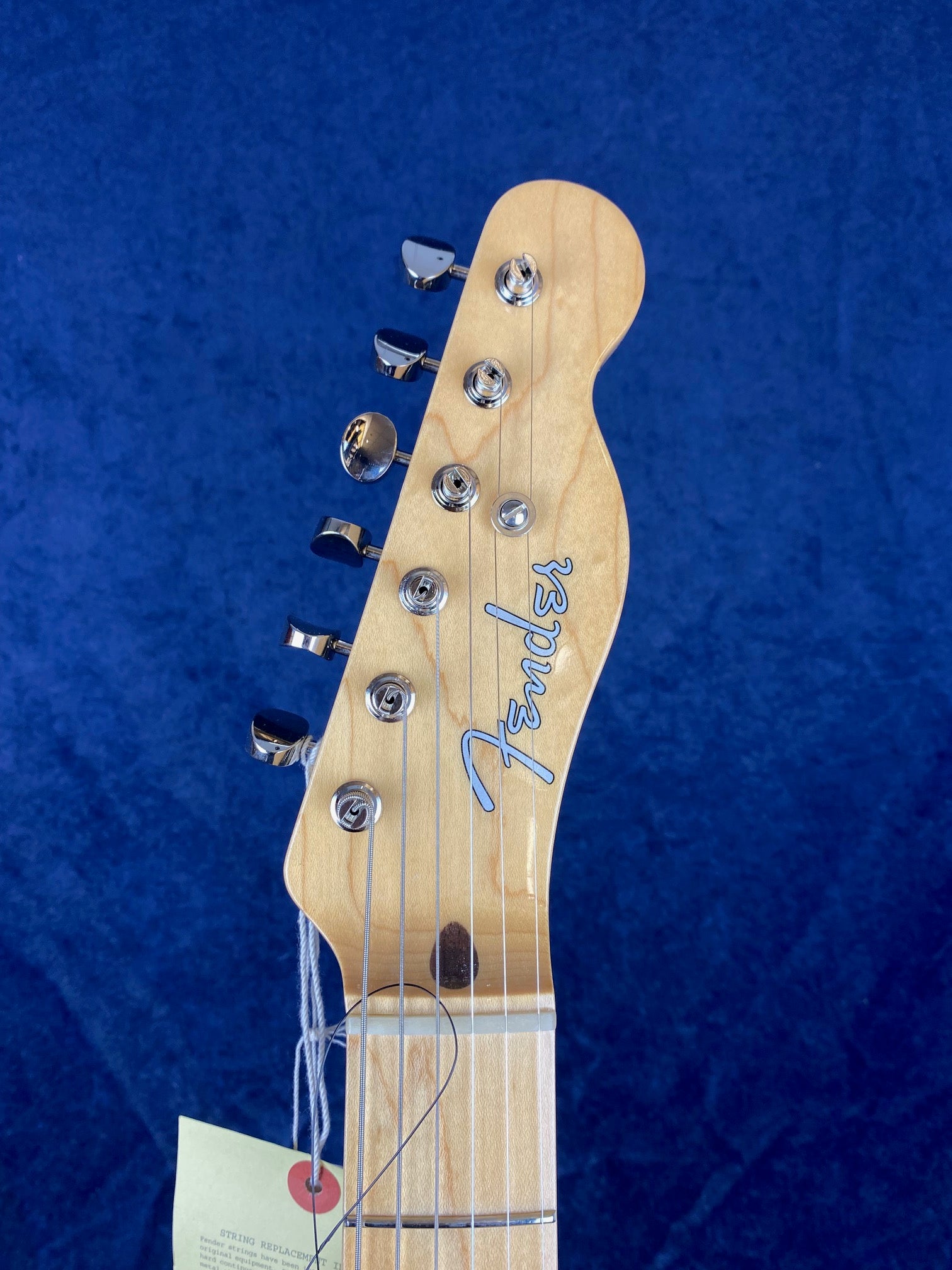 Maple Fender guitar headstock with six chrome tuning pegs and the Fender logo, set against a deep blue background. A tag hangs from a string.