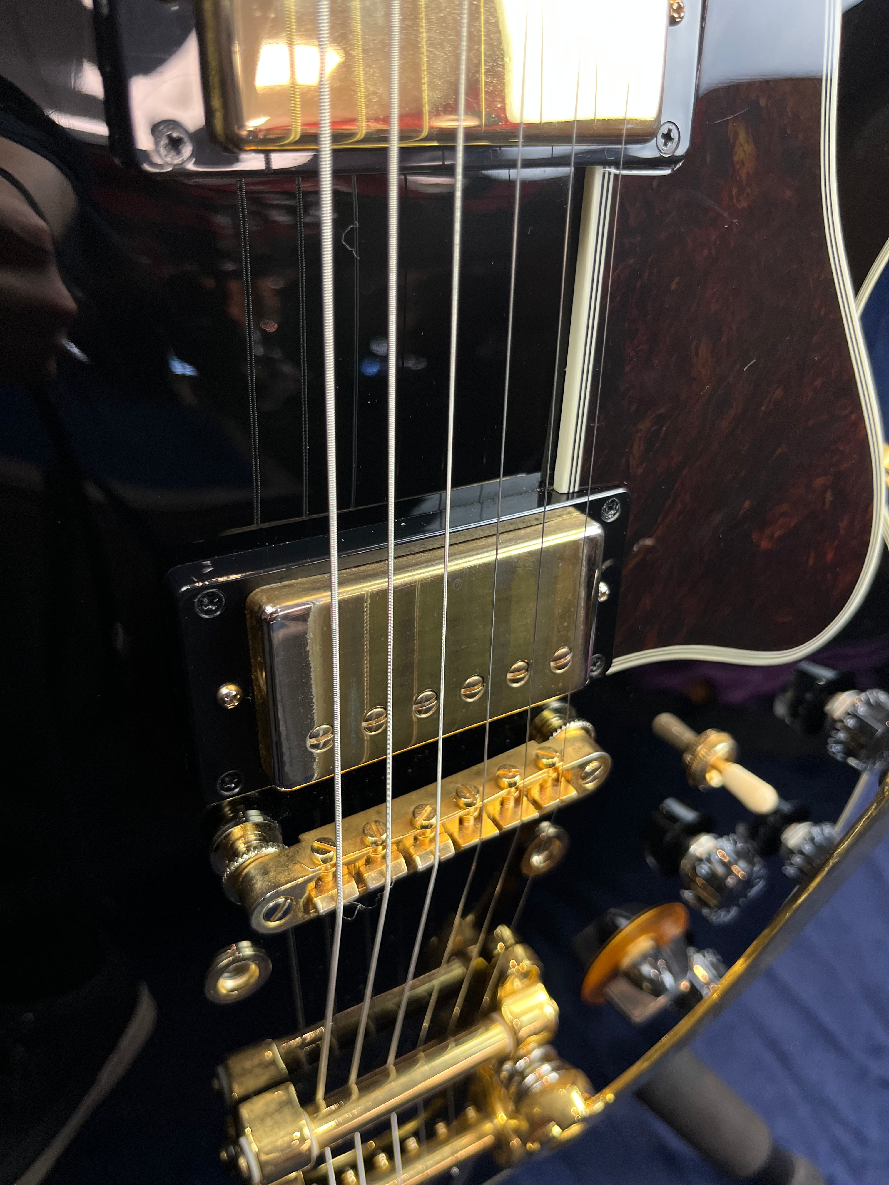 Close-up of an electric guitar showing gold humbucker pickup, gold bridge, vibrato system, black knobs, and dark glossy body with cream binding.