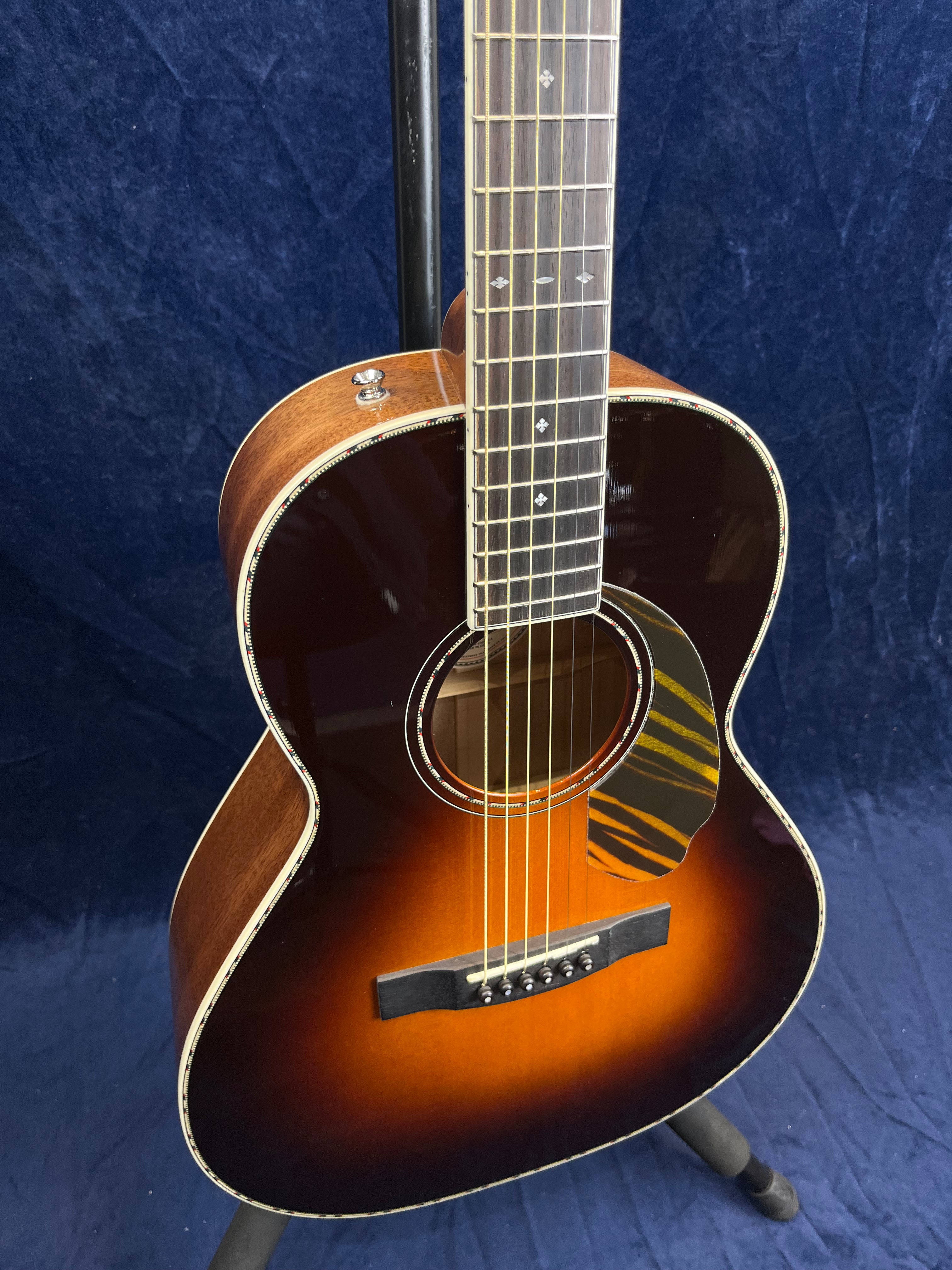 Sunburst finish acoustic guitar with a decorative pickguard, pearl fretboard inlays, and cream binding, photographed against a dark blue backdrop.
