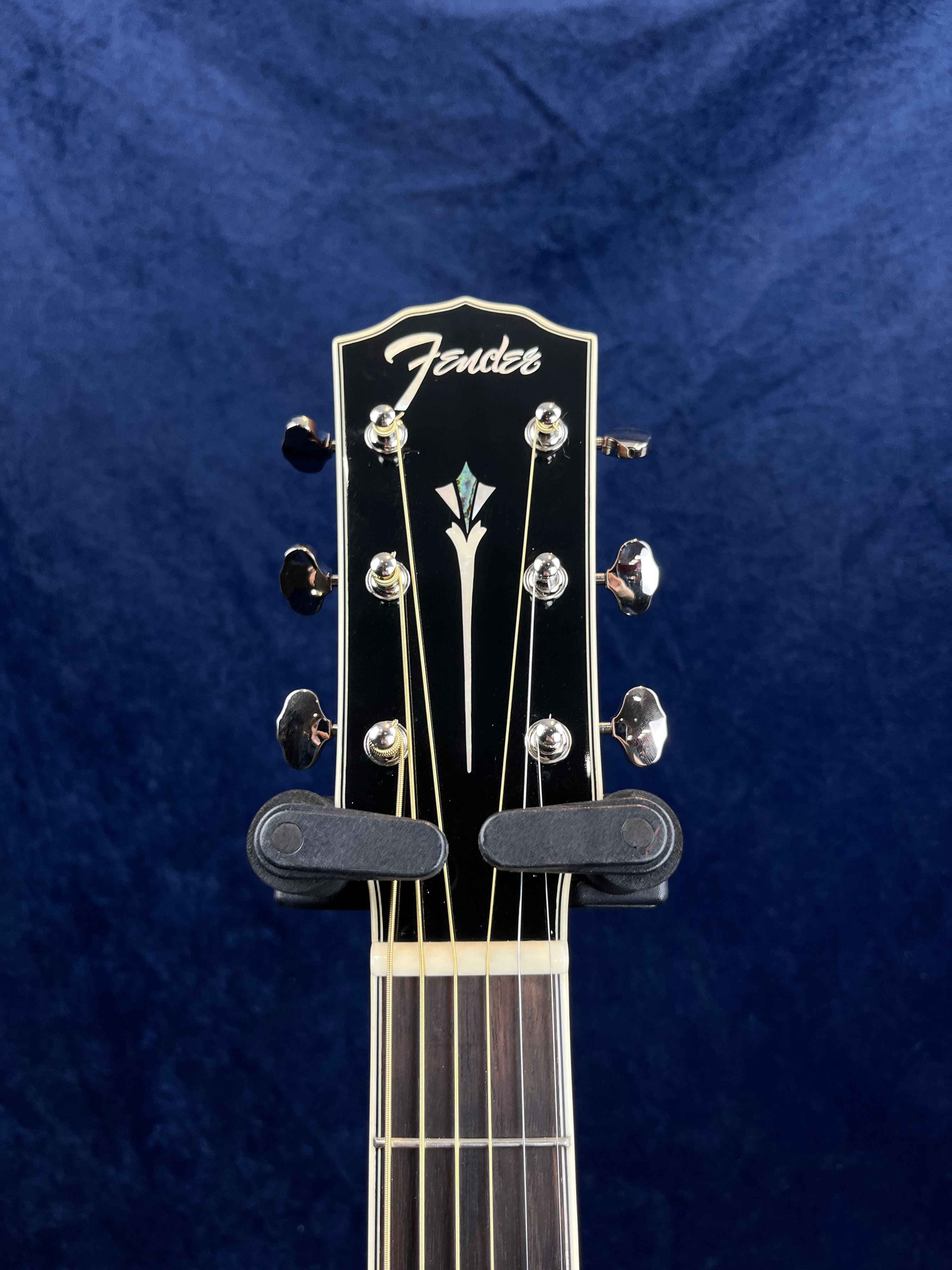 The headstock of a Fender acoustic guitar with six silver tuning pegs and decorative inlay, held by a black guitar hanger against a dark blue background.