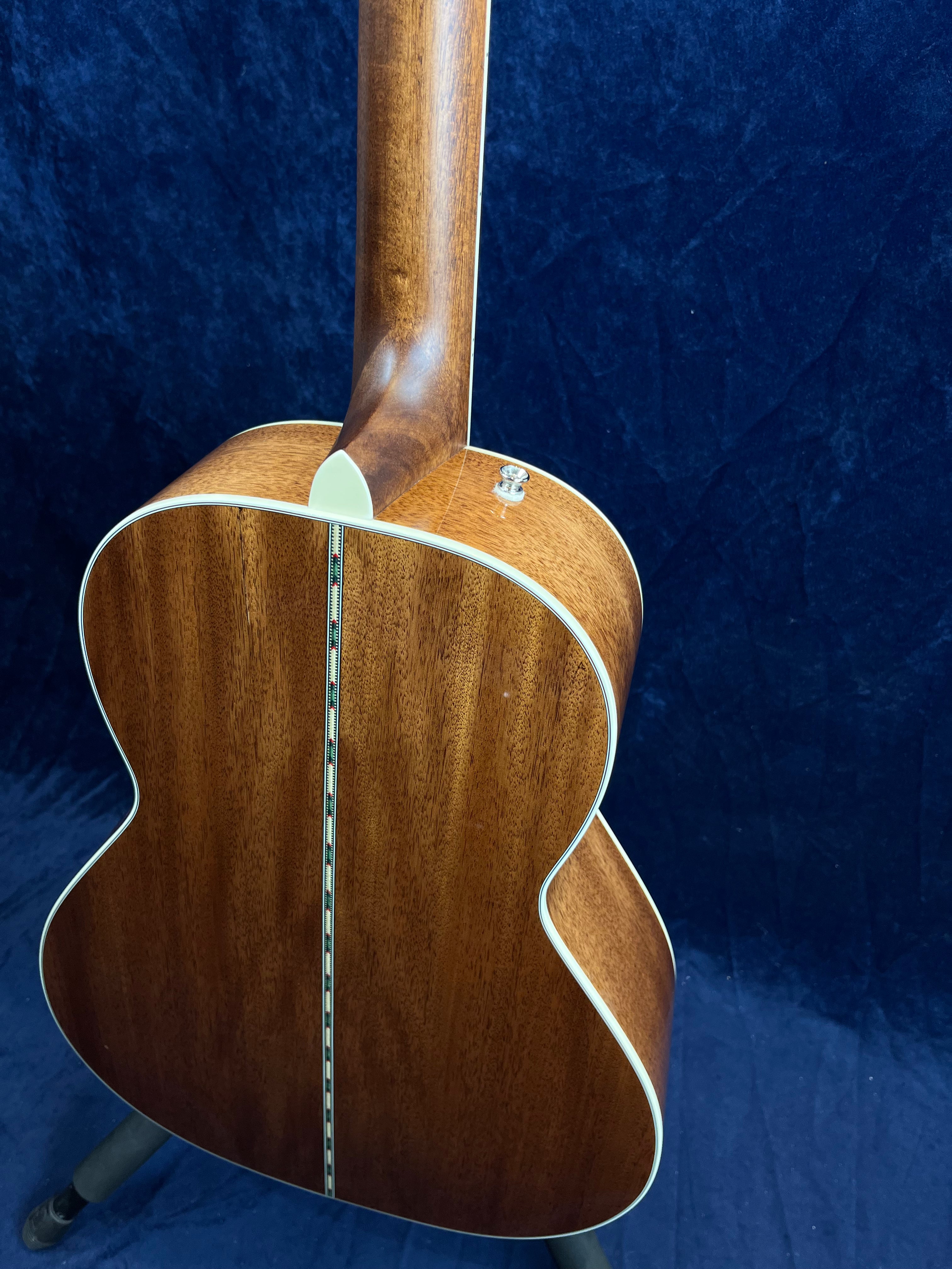 Brown acoustic guitar back and neck with cream binding, a decorative center strip runs down the back, resting on a stand against a dark blue background.