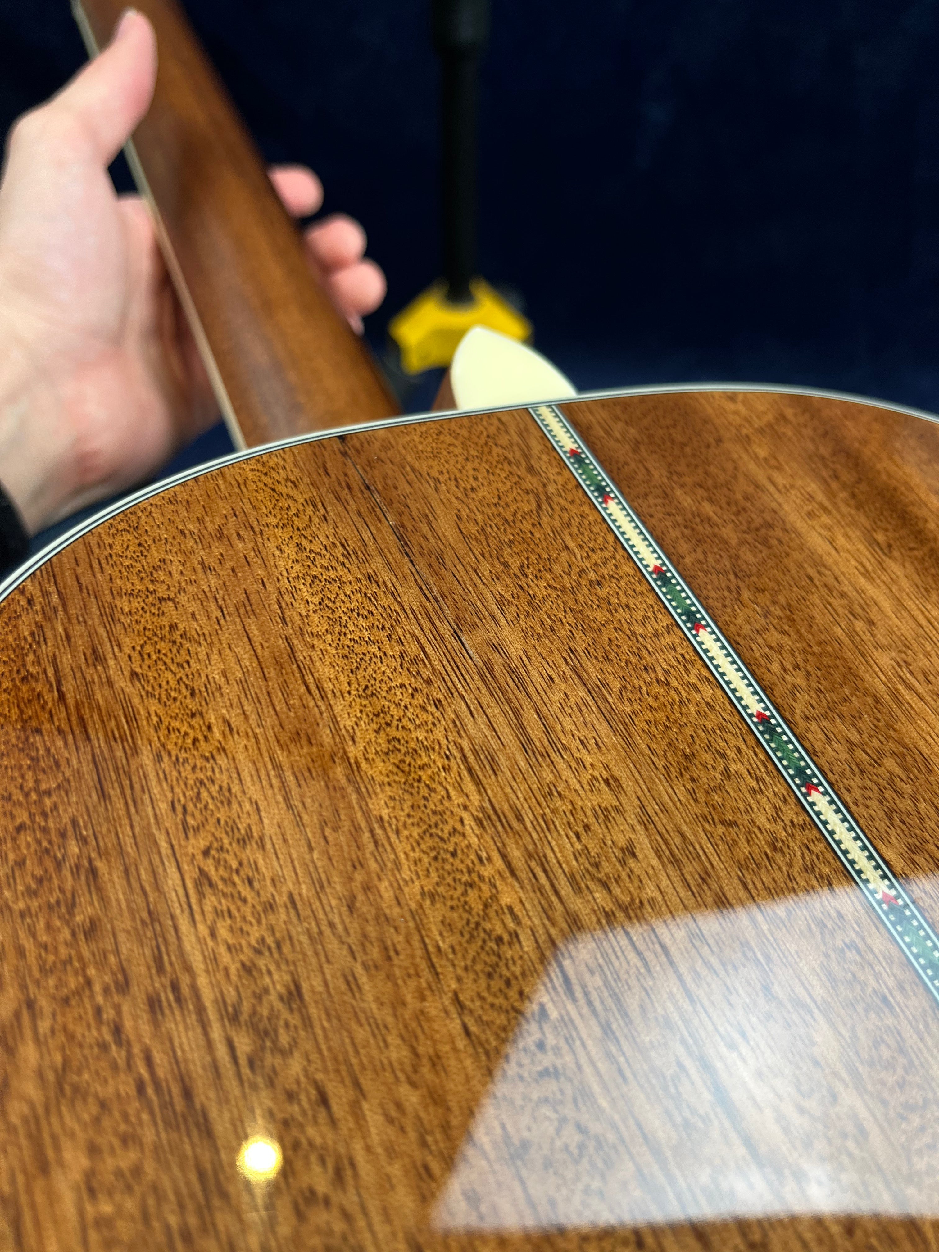 Hand holding an acoustic guitar with a glossy wood finish; back has a decorative inlay strip running down the center.