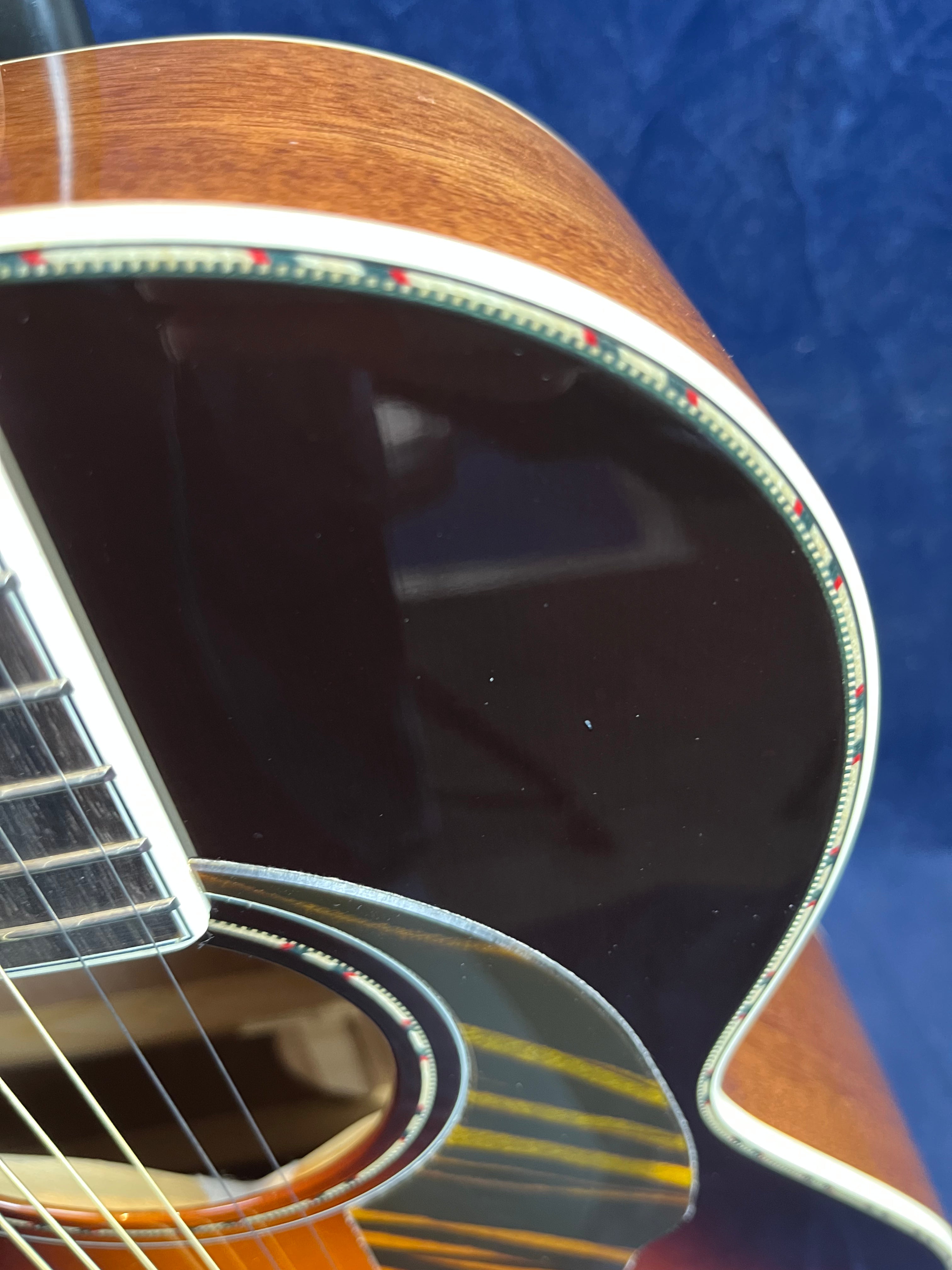 Close-up of an acoustic guitar body showing brown wood, black pickguard, detailed soundhole rosette, and cream-colored binding with red accents.