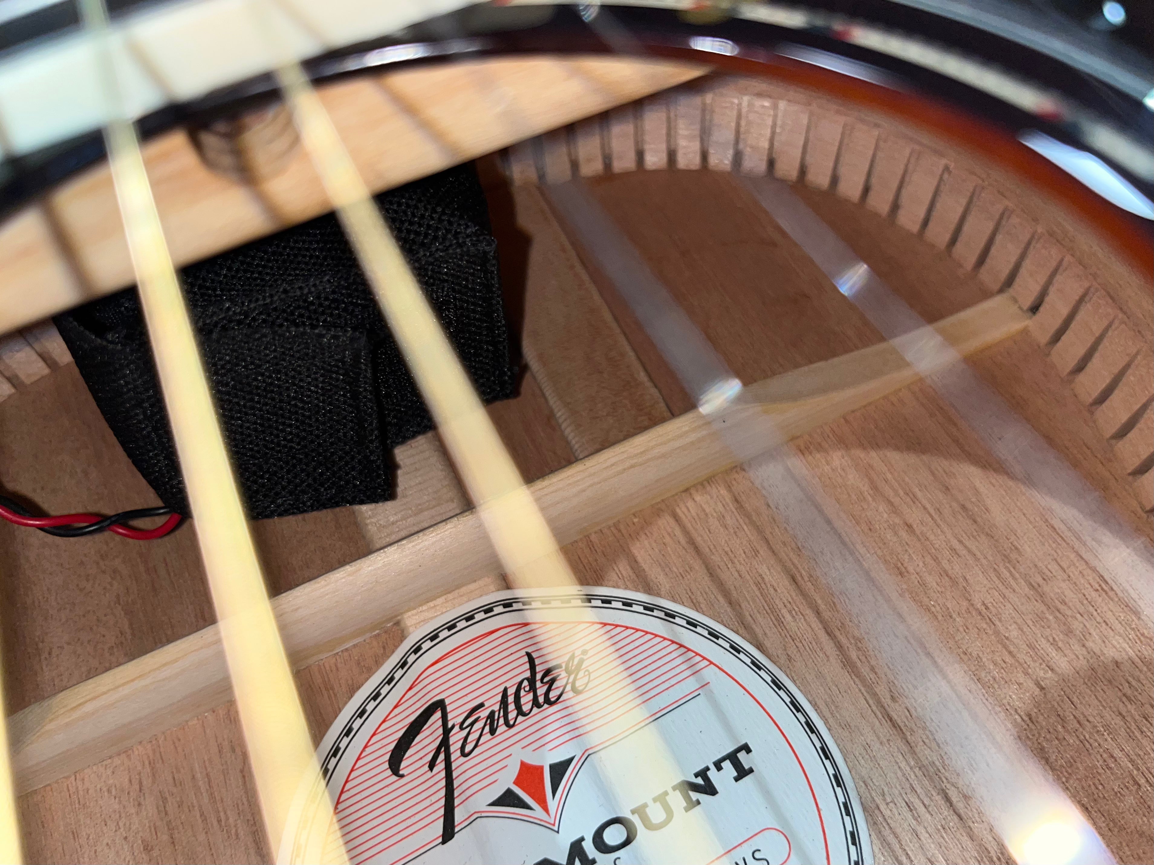 View inside an acoustic guitar shows Fender label, wooden bracing, nylon strings, and a small black pouch with red and black wires attached.