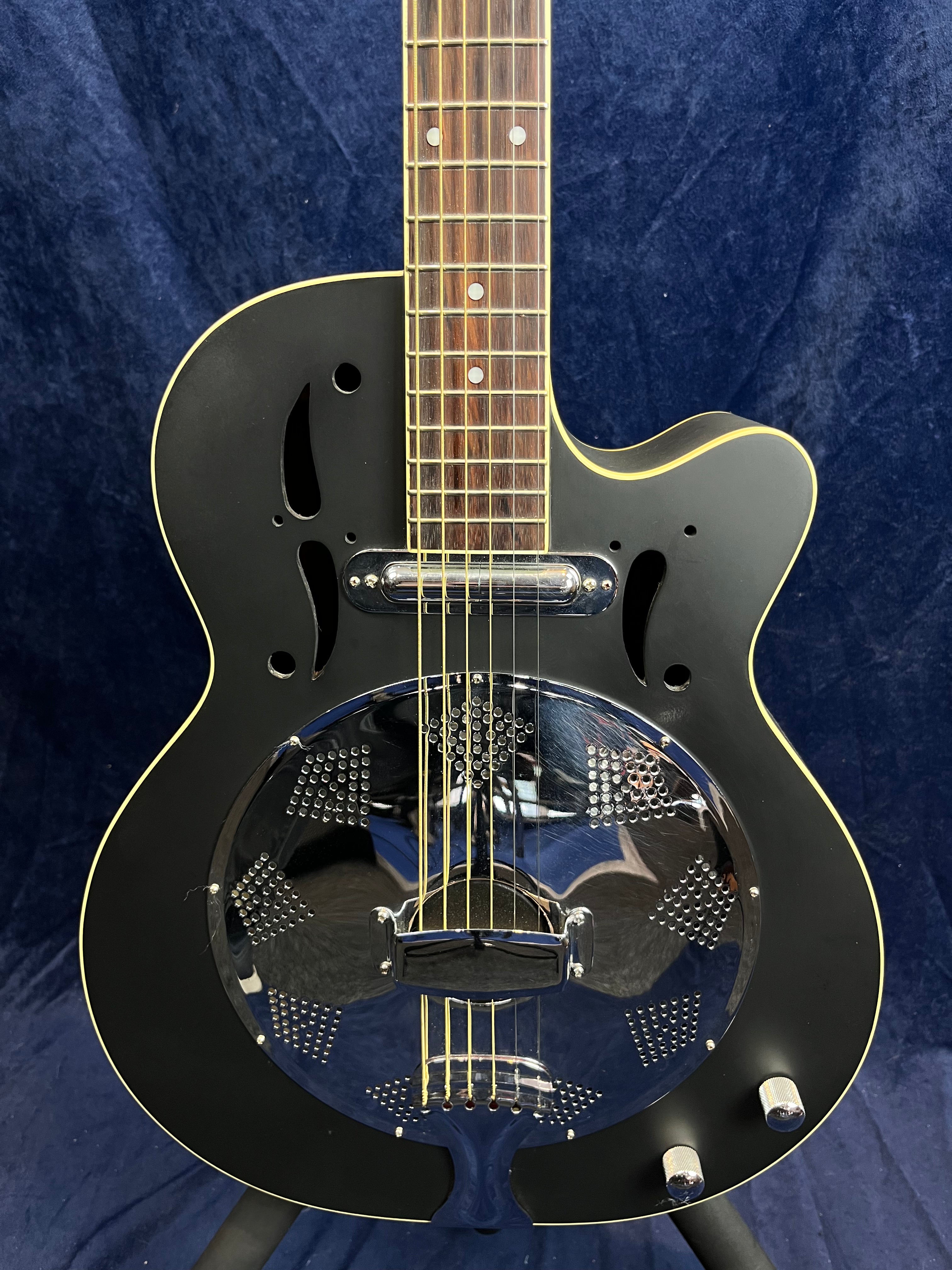 Guitare électrique noire à corps simple pan coupé avec résonateur métallique rond et boutons de réglage argentés en bas.